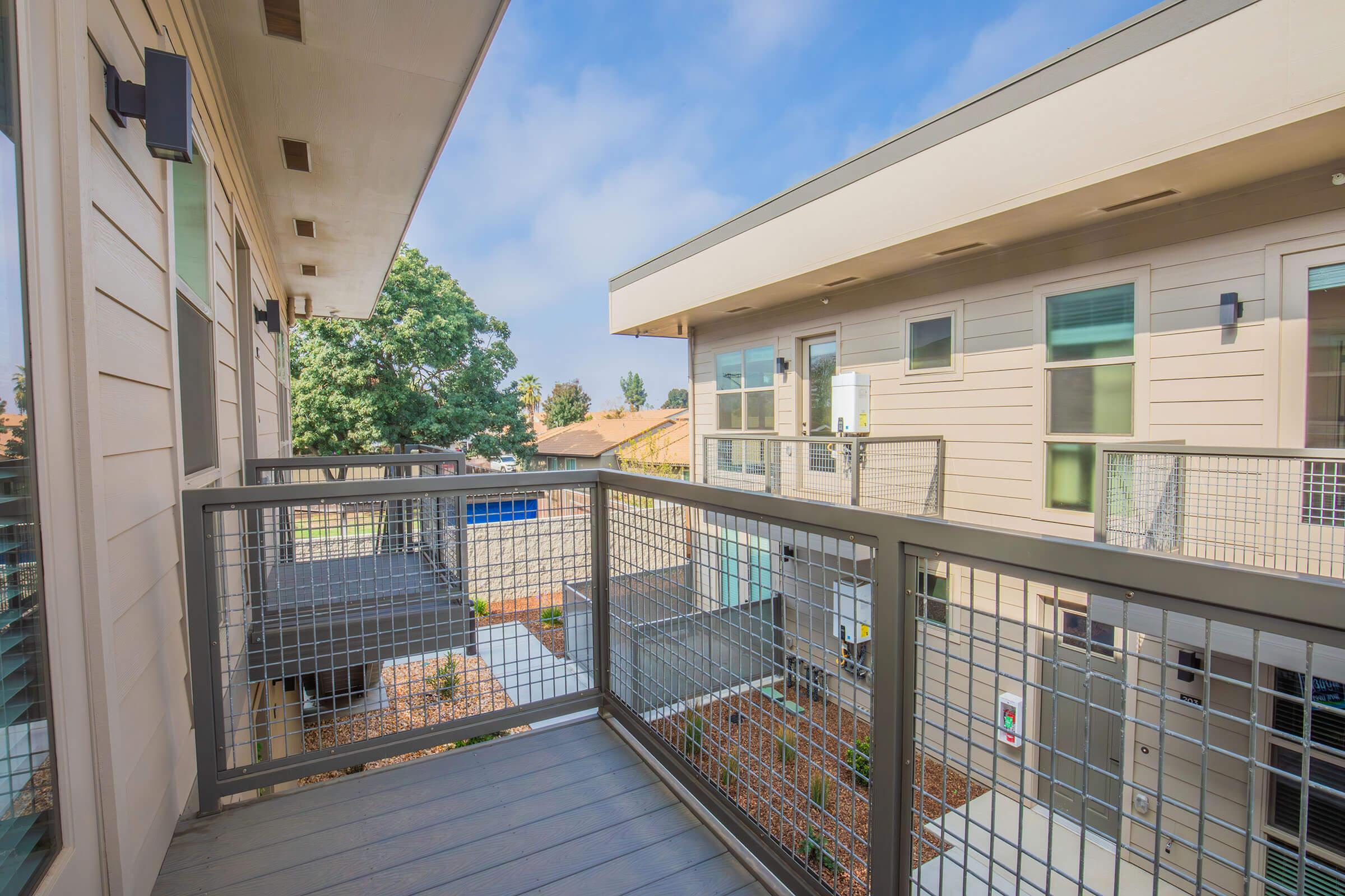 A modern apartment balcony overlooking a courtyard, with a gray railing and wooden flooring. The view includes nearby trees and a well-maintained landscape. The buildings feature contemporary architecture with large windows. The sky is partly cloudy.