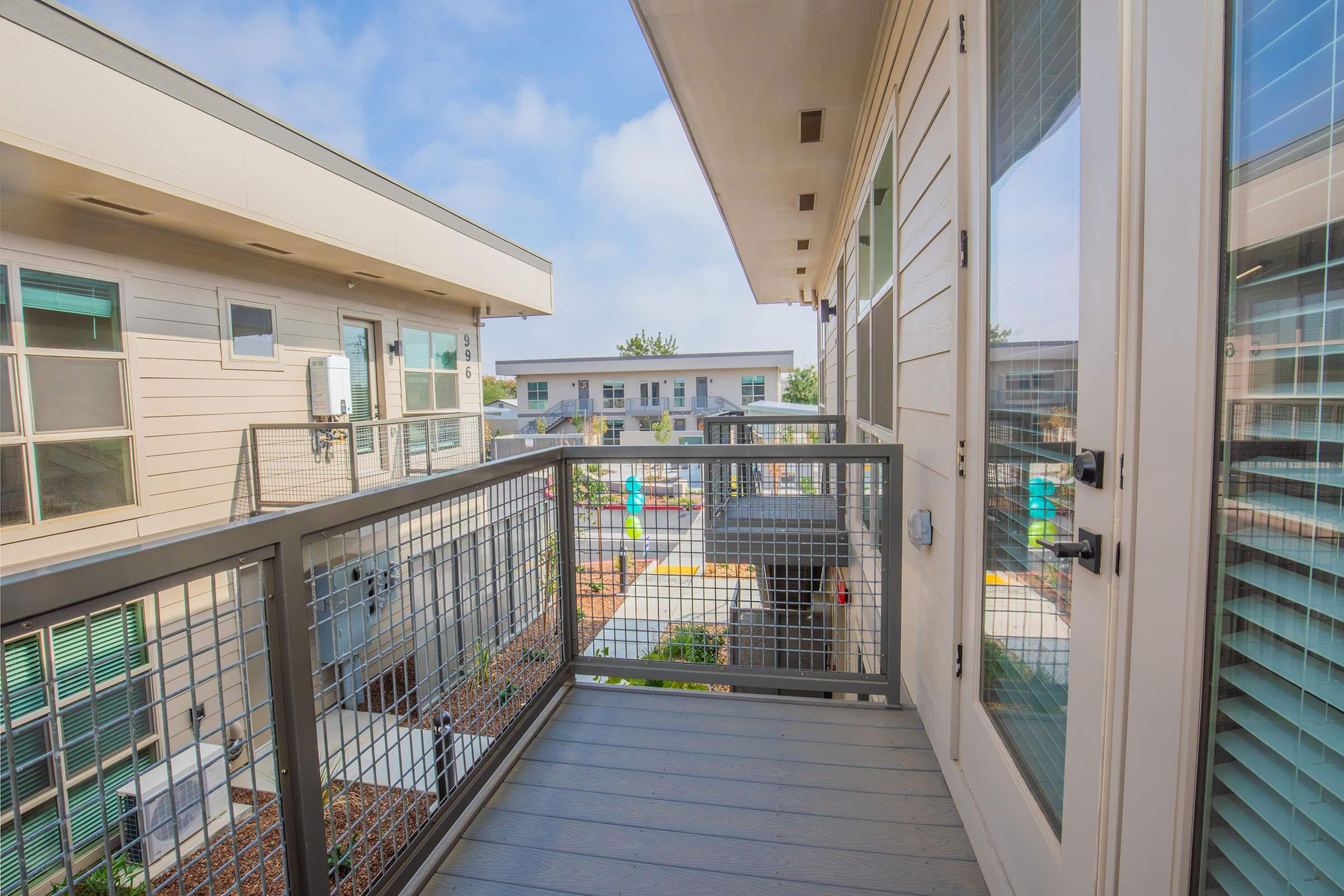 A view from a balcony overlooking a modern residential complex. The balcony features a railing and is surrounded by buildings with large windows. In the background, there are colorful outdoor play areas, trees, and a clear blue sky, creating a bright and inviting atmosphere.