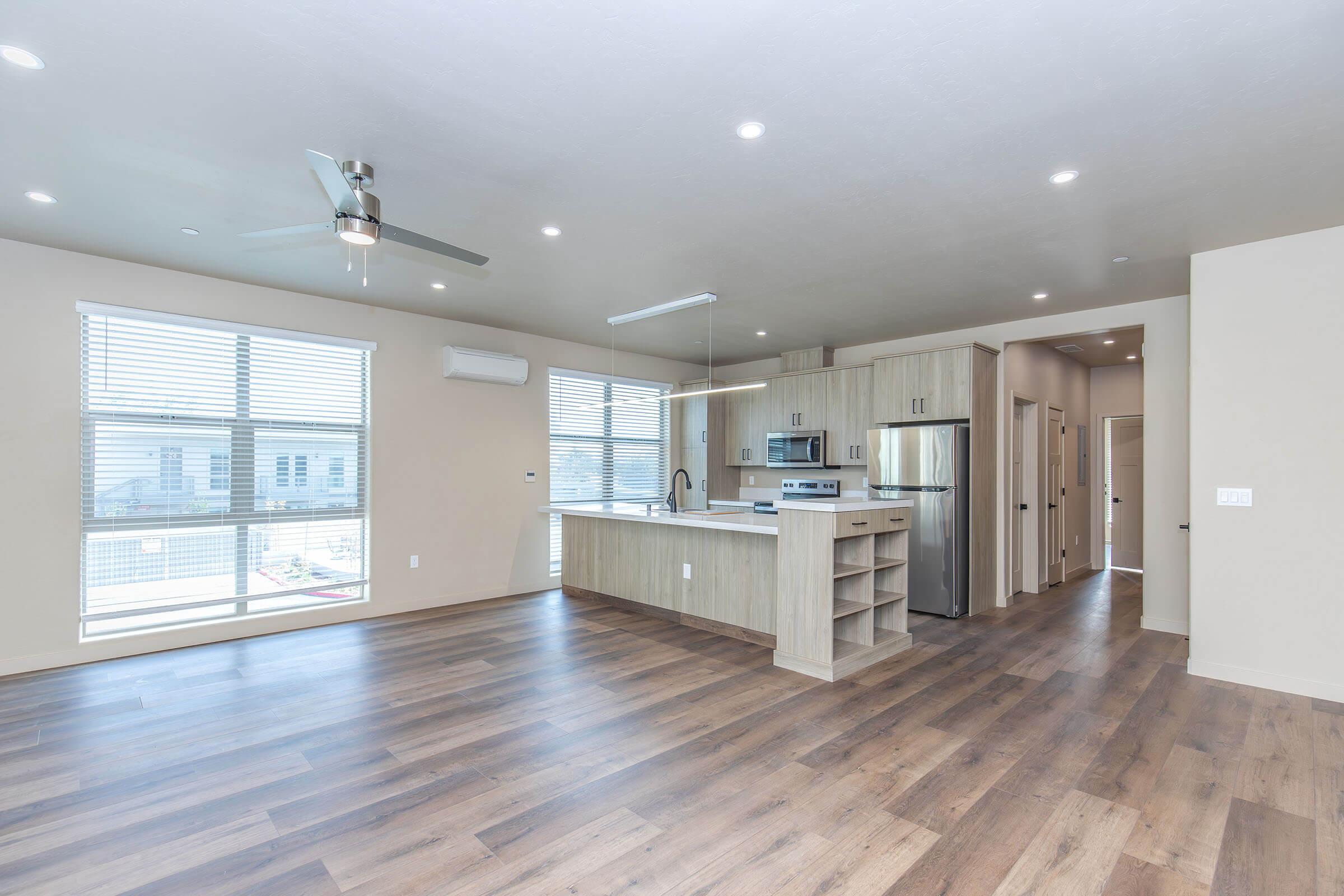 Modern open-concept living space featuring hardwood floors, large windows with blinds, and a well-equipped kitchen with stainless steel appliances and a central island. The area is brightly lit with recessed ceiling lights and a ceiling fan, providing a spacious and inviting atmosphere.
