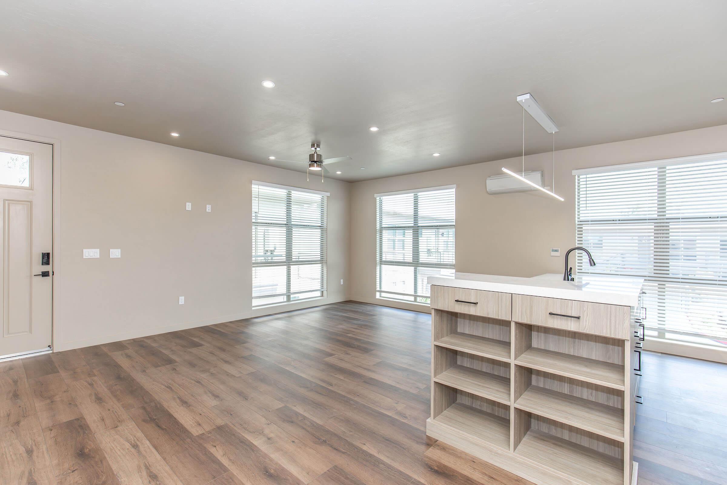 An open-concept living space featuring large windows, neutral-colored walls, and wooden flooring. There is a contemporary kitchen island with shelving, a modern light fixture above, and minimal furnishings. Natural light brightens the room, creating a spacious and inviting atmosphere.