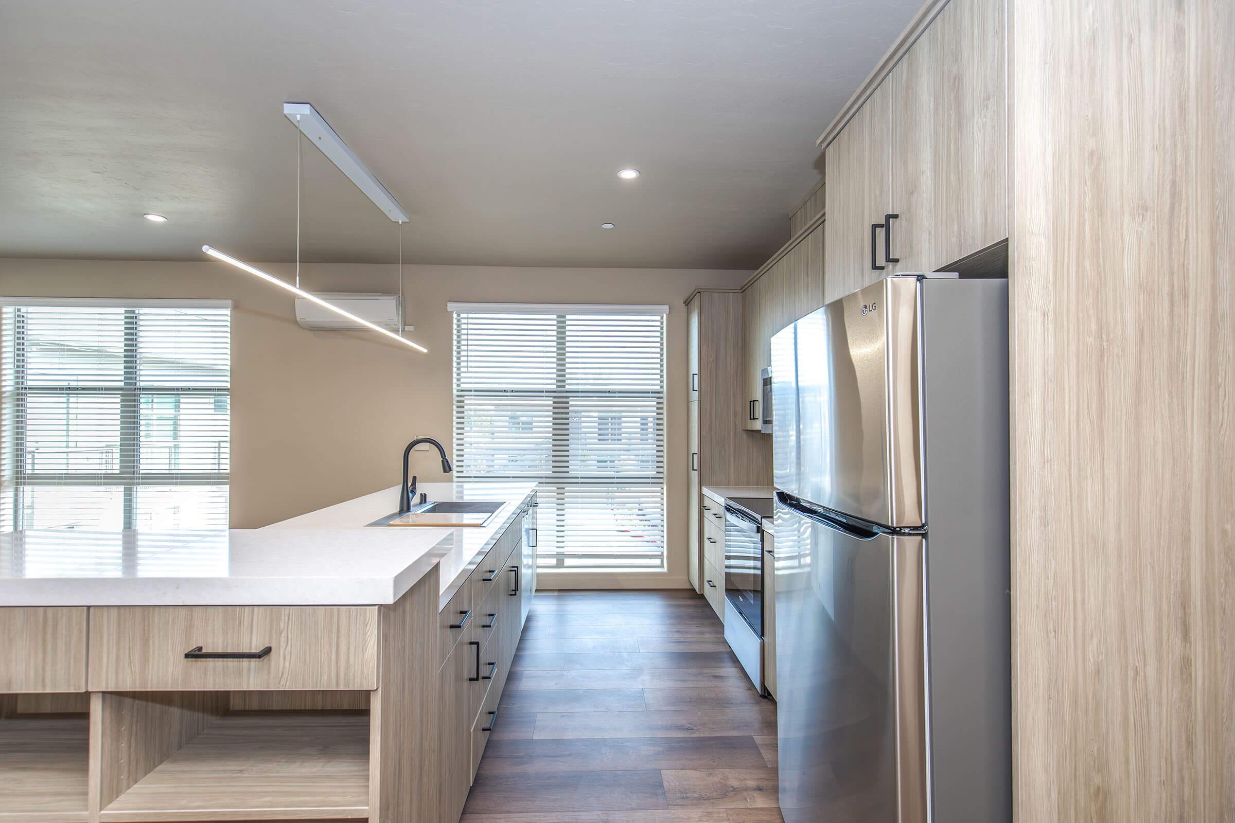 Modern kitchen featuring light wood cabinetry, stainless steel appliances, and a sleek white countertop. There is a large window with blinds allowing natural light, and an island bar with a sink and minimalist pendant lighting above. The flooring is dark wood, creating a warm contrast with the lighter cabinets.
