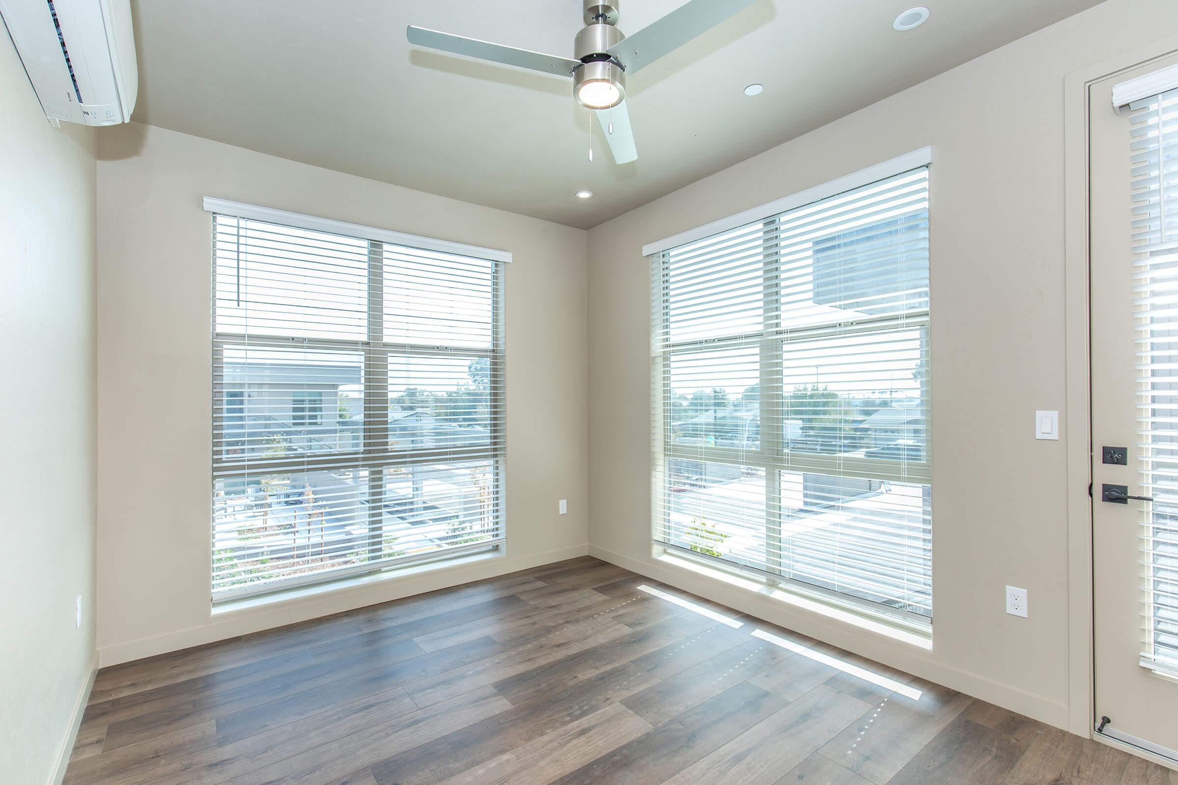 Bright, modern room with light-colored walls and wooden flooring. Two large windows with blinds provide natural light, and a ceiling fan is mounted in the center. A door leads outside, and the overall feel is spacious and airy. Ideal for a living space or office.