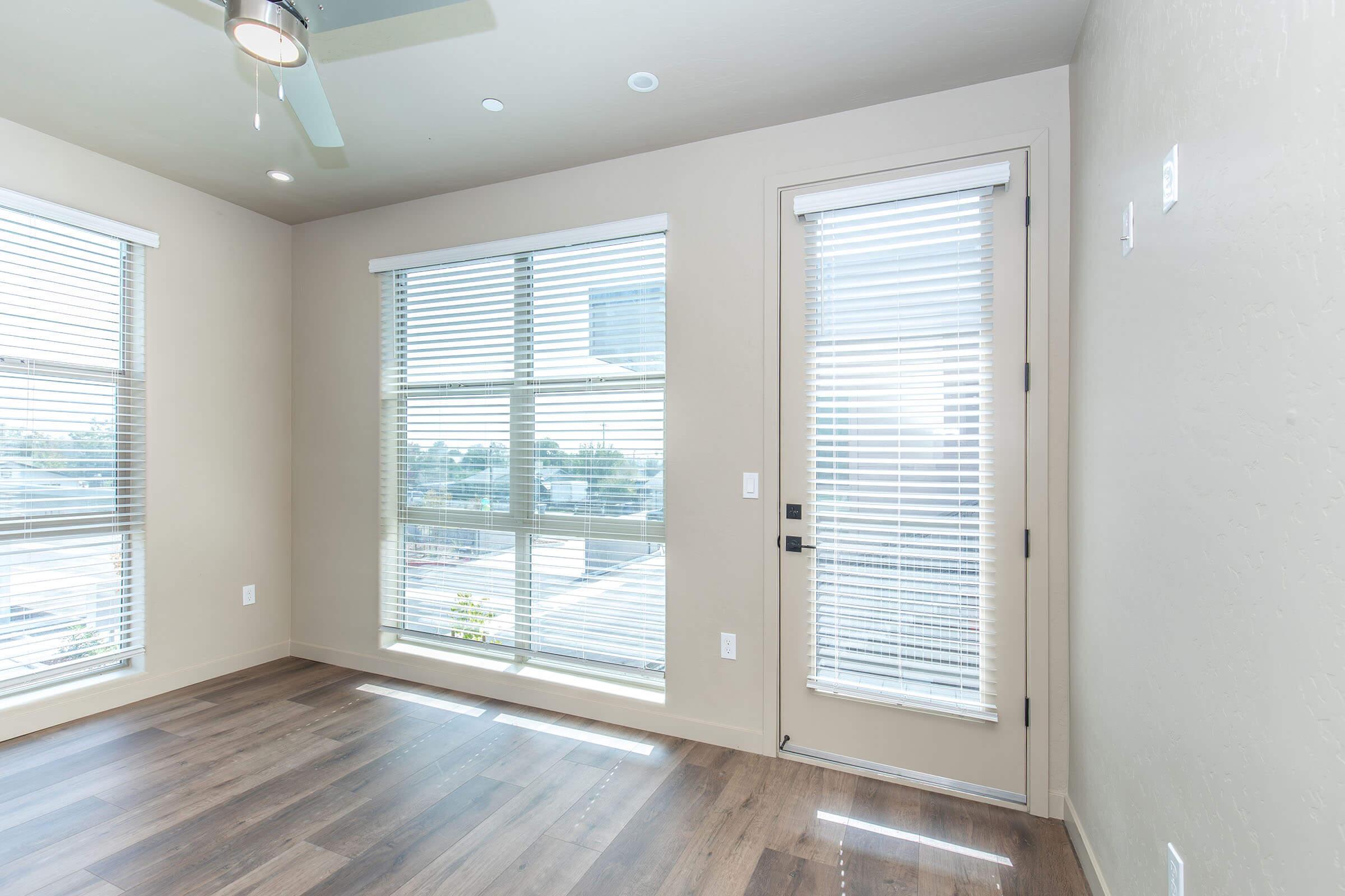 Bright, modern room with large windows featuring white blinds, hardwood flooring, and light-colored walls. A ceiling fan is visible, and a door with a window leads outside. The space has a clean and airy feel, ideal for natural light.