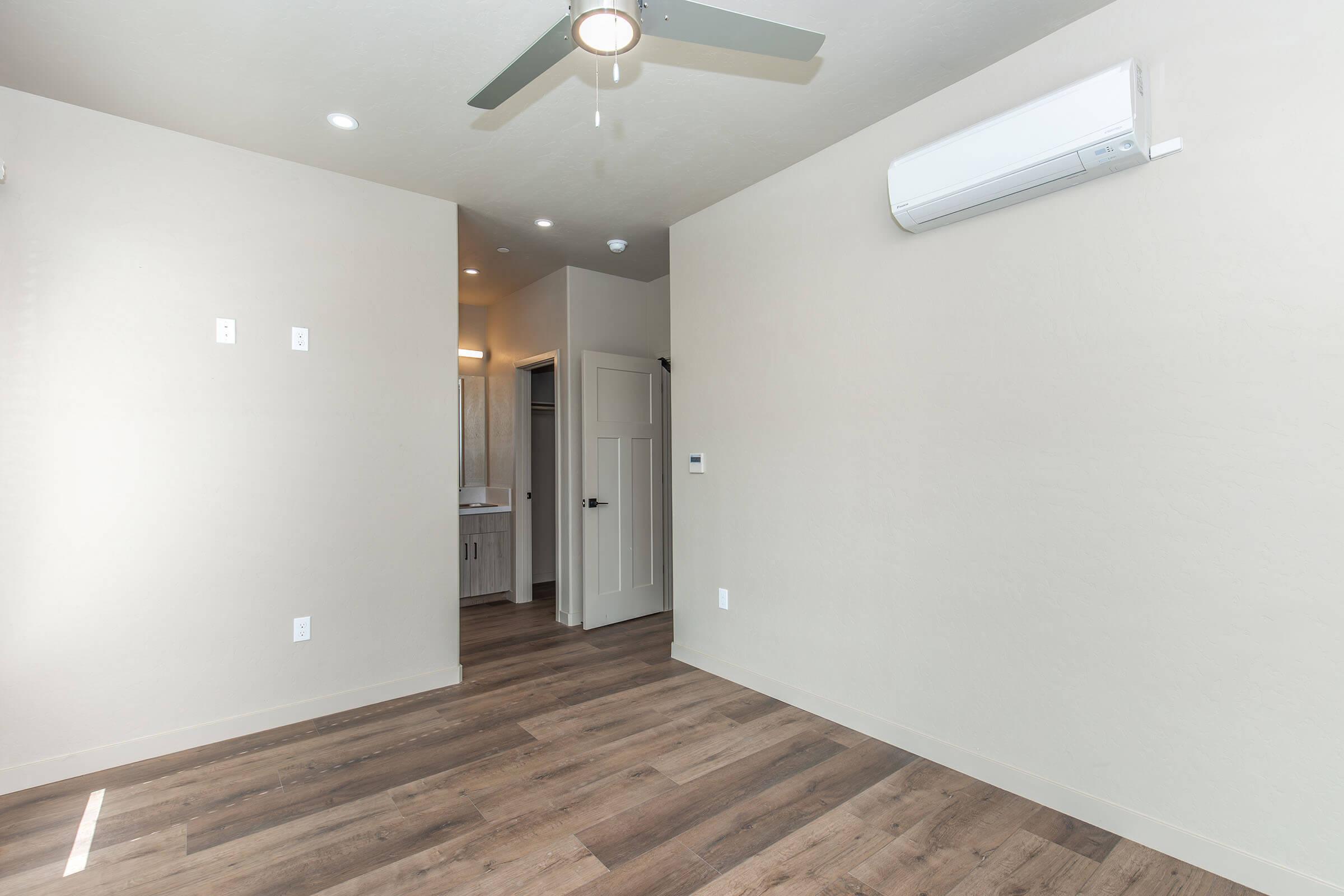 A bright, empty room featuring light-colored walls, a ceiling fan, and wooden flooring. There is an air conditioning unit mounted on the wall and a door leading to a bathroom or closet in the background, creating a spacious and modern atmosphere.