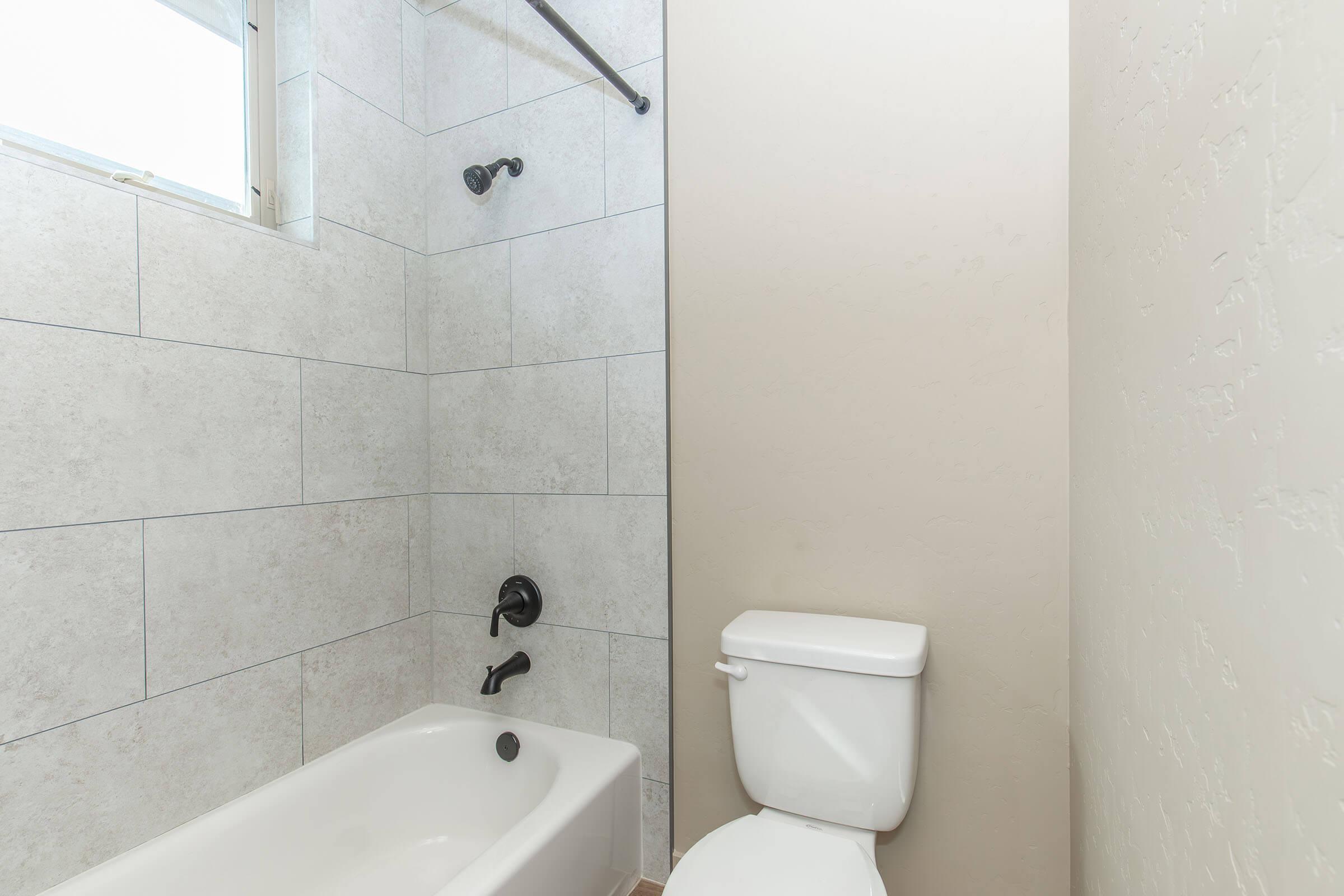 A bathroom with a bathtub and shower combination, featuring light-colored tiled walls. A black showerhead and faucet are mounted on the wall, and a white toilet is positioned beside the bathtub. A window provides natural light to the space.