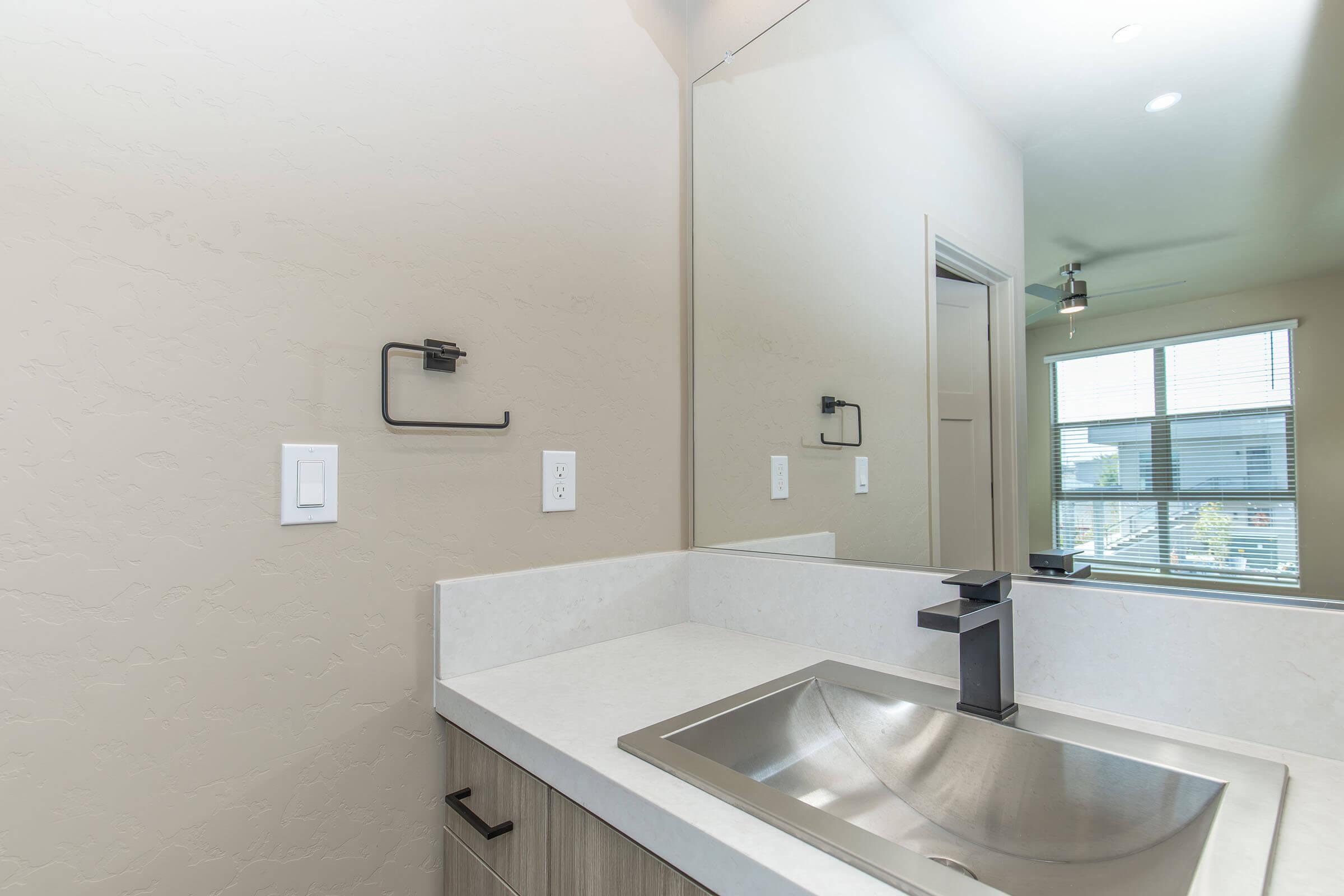 A modern bathroom sink area featuring a sleek stainless steel sink, simple countertop, and a large wall mirror. The wall is painted in a light neutral color, with a towel holder and light switch visible. Natural light filters through a window, enhancing the contemporary design.