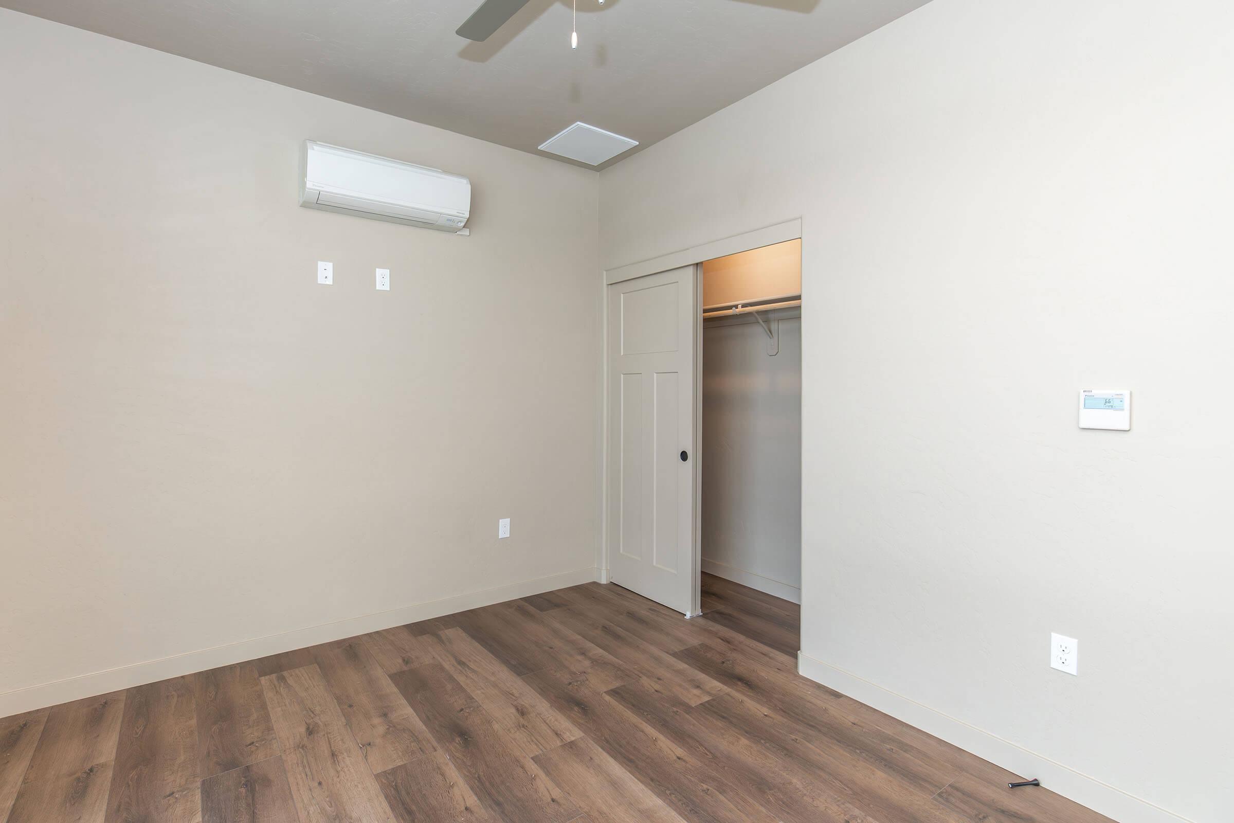 A clean, empty room with light-colored walls and wooden flooring. It features a ceiling fan, an air conditioning unit, and a closet with sliding doors. The room has minimal furnishings, creating a spacious feel. Natural light may filter in through the open door and windows.