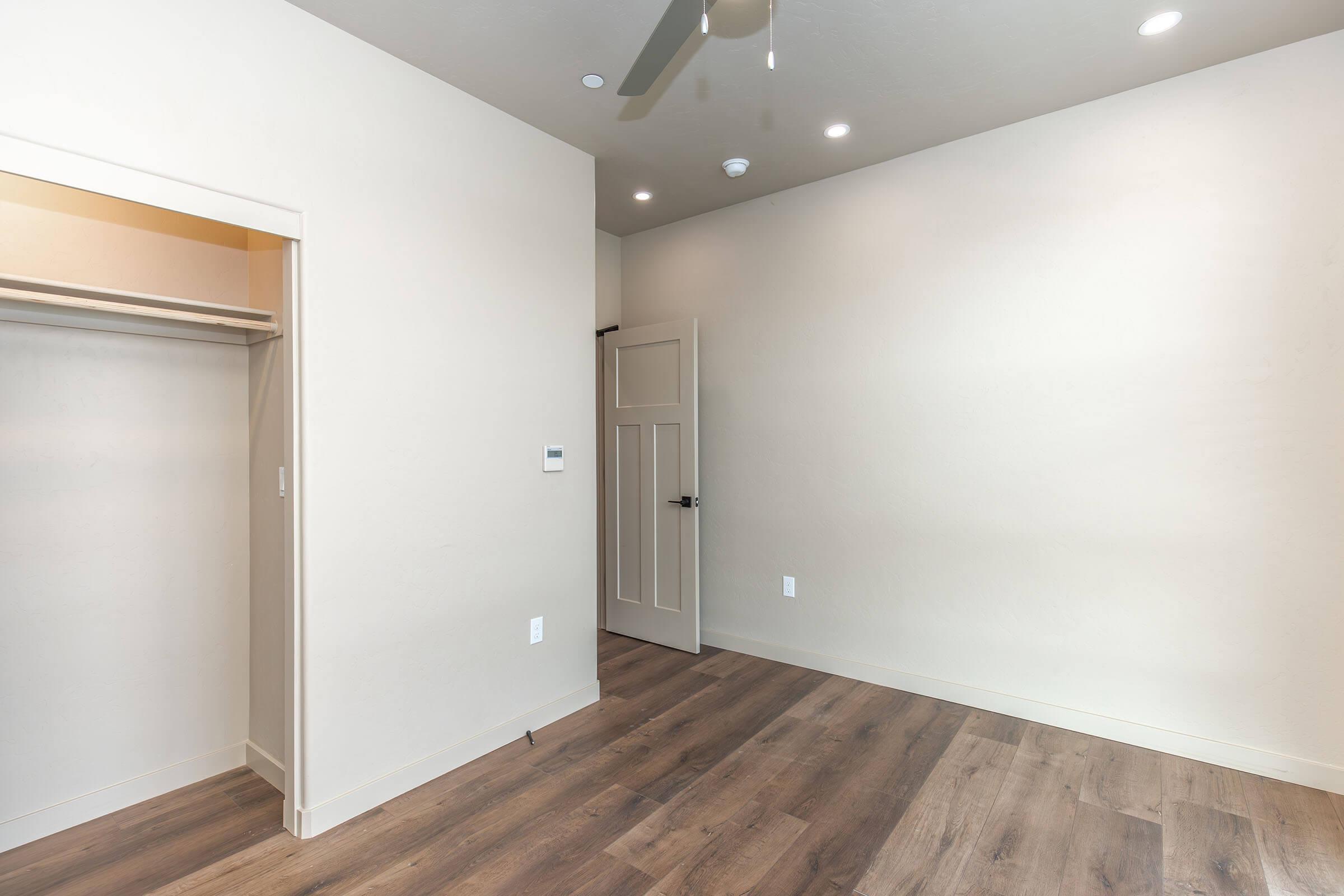 Interior view of a room featuring light-colored walls, a ceiling fan, and recessed lighting. There is an open closet on the left with a simple door, while the opposite wall has a closed door leading to another space. The floor is a warm wood tone, contributing to a modern and minimalist aesthetic.