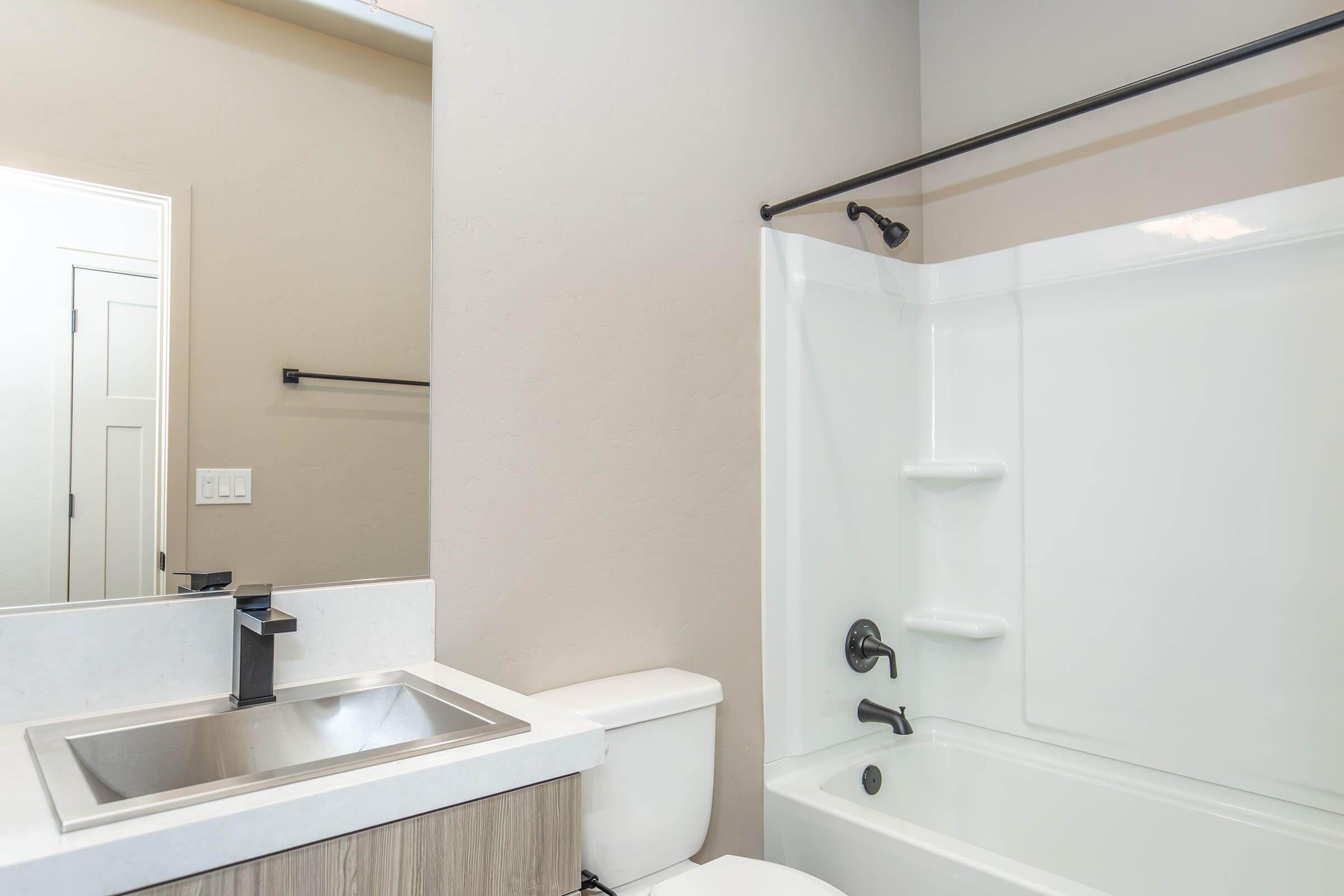 A modern bathroom featuring a white bathtub with a shower, a sleek sink with a black faucet, and a toilet. The walls are painted in a neutral color, and there is a mirror above the sink. A towel bar is mounted on the wall, and a door is visible in the background.