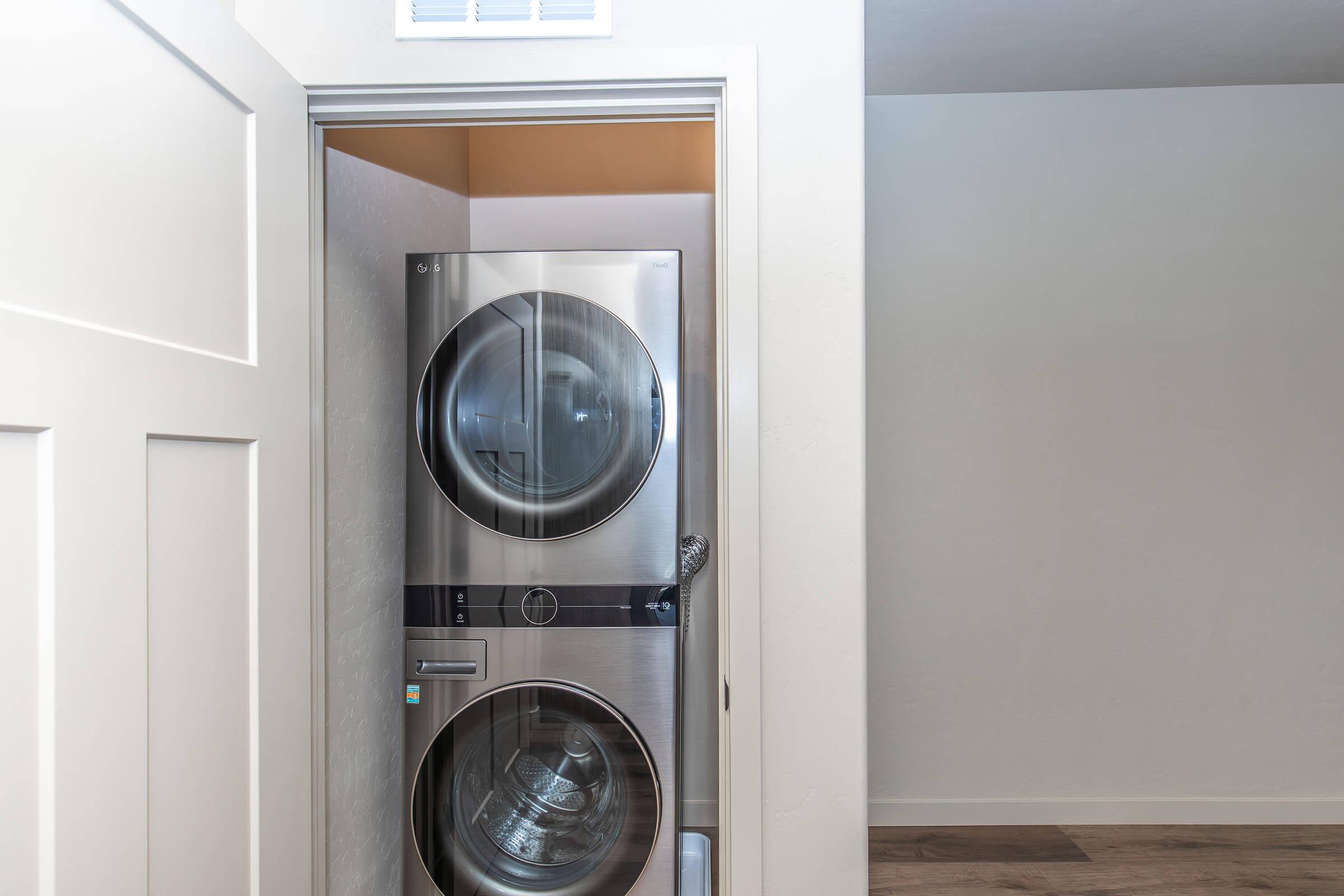A compact laundry area featuring a stacked washer and dryer in stainless steel, placed within a small alcove. The surrounding walls are a light color, and the room has a minimalist, modern design with wooden flooring. A partially open door leads to the laundry space.