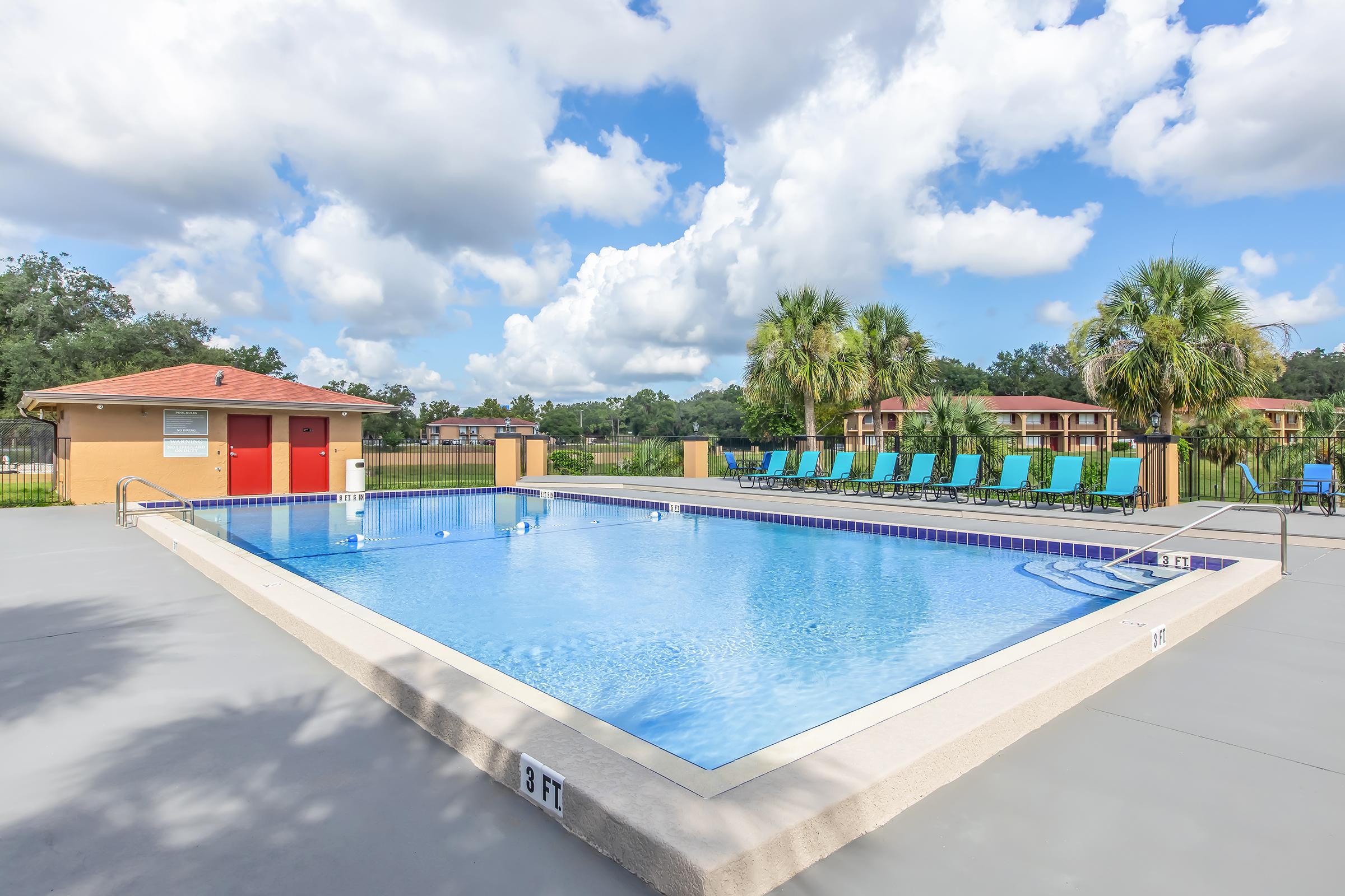 A clear blue swimming pool with lounge chairs arranged around it, located in a sunny outdoor area. A poolside building with red doors is visible in the background, along with palm trees and suburban buildings under a partly cloudy sky. The pool depth is marked with a "3 FT" sign.