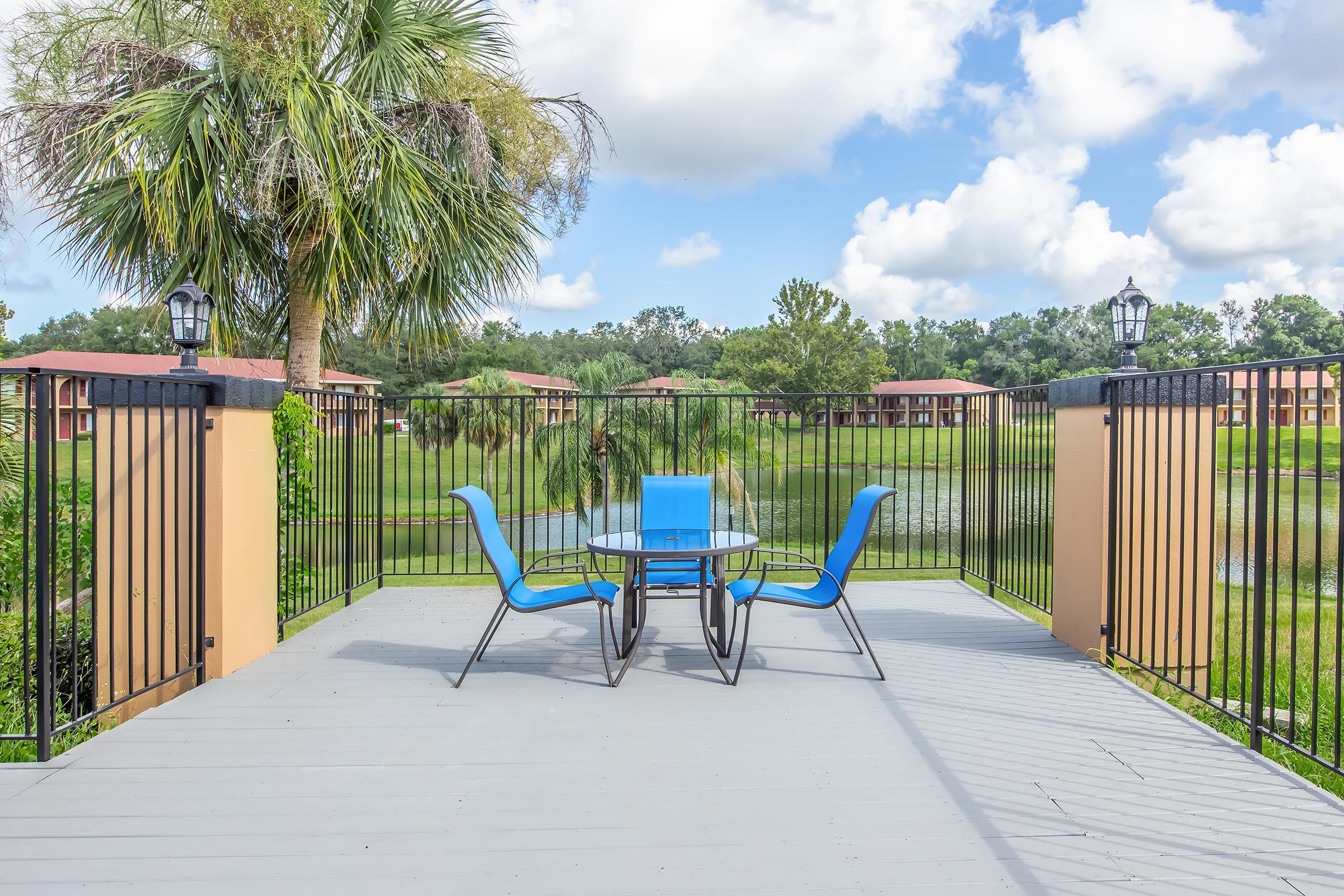 A wooden deck overlooking a serene pond, featuring a glass table surrounded by blue chairs. Lush greenery and palm trees provide a tropical ambiance, with residential buildings visible in the background under a partly cloudy sky.
