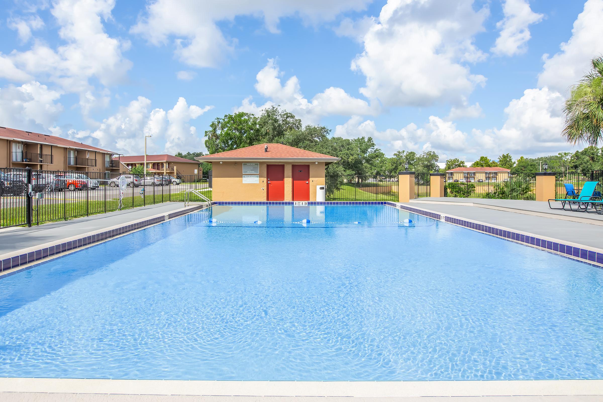 A spacious outdoor swimming pool with clear blue water, surrounded by a concrete deck. In the background, there are green lawns and residential buildings. A small pool house with red doors is visible beside the pool, under a bright sky filled with fluffy white clouds.