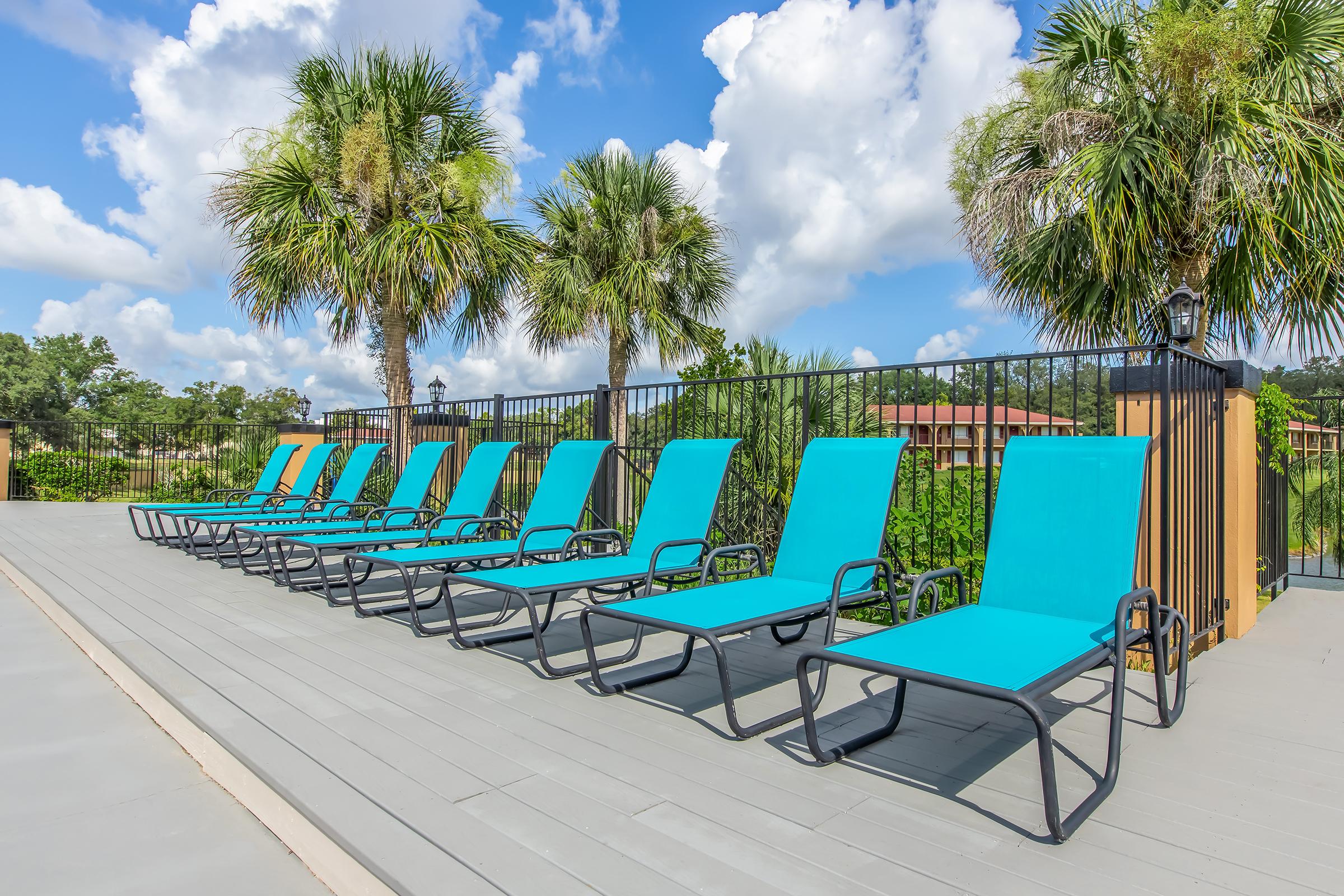 A row of vibrant turquoise lounge chairs arranged neatly on a wooden deck, surrounded by lush greenery and palm trees. The sky is bright blue with fluffy white clouds, creating a sunny and inviting atmosphere. Ideal for relaxation and enjoying outdoor spaces.