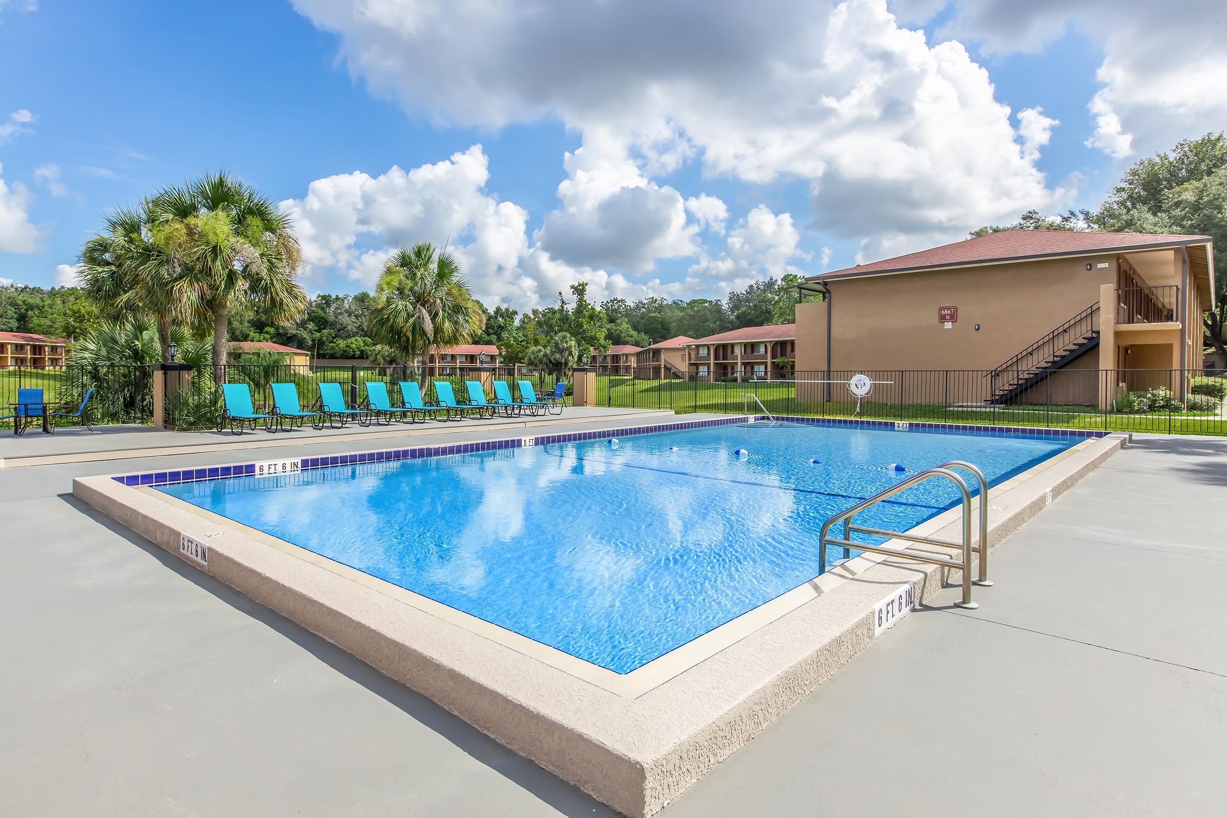 A clear blue swimming pool surrounded by lounge chairs under a bright sky with fluffy clouds. In the background, a two-story building is visible, along with lush greenery and palm trees. The pool area is well-maintained and inviting, creating a relaxing atmosphere.