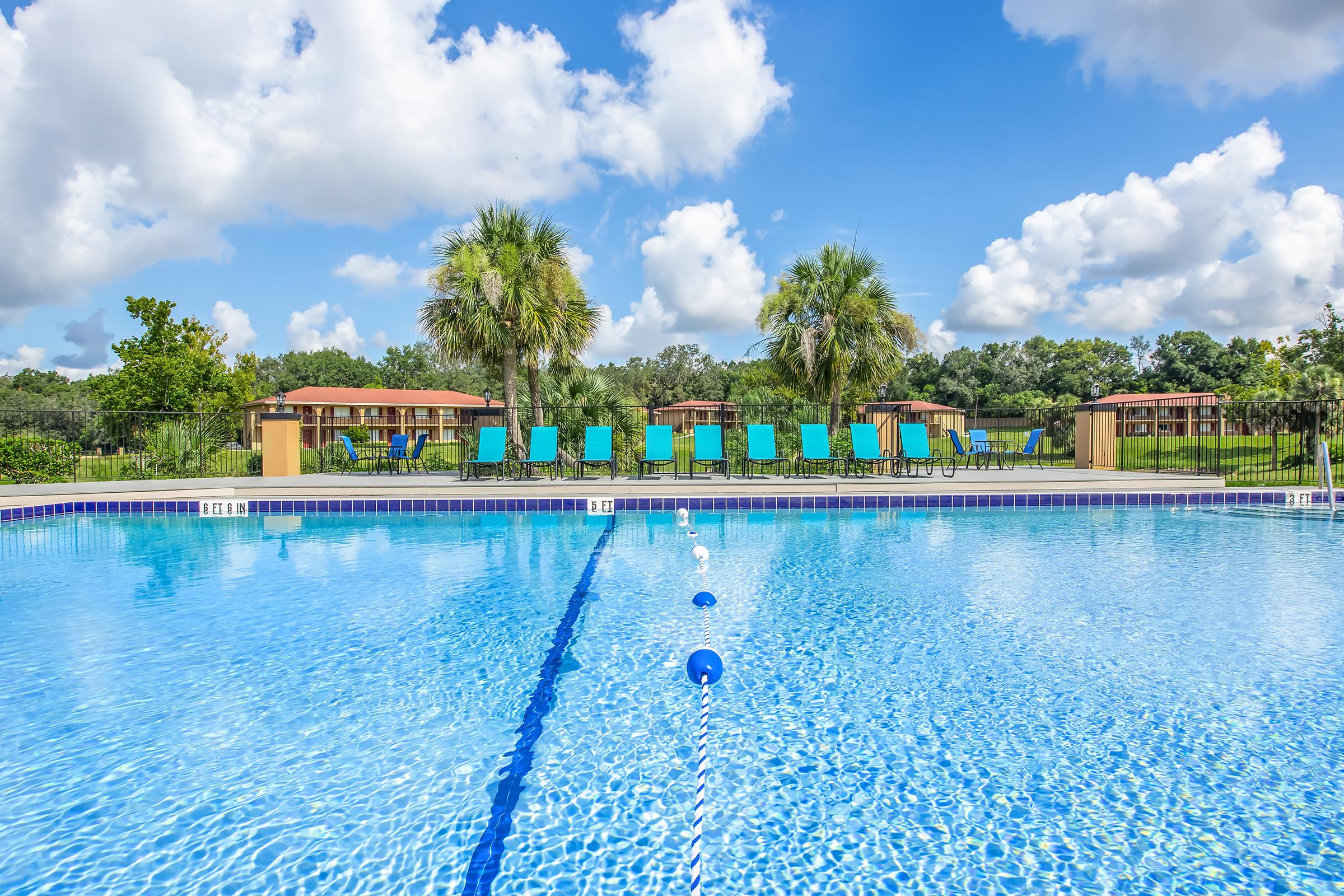 A clear swimming pool with a bright blue surface, surrounded by lush greenery and palm trees. In the background, there are several turquoise lounge chairs and cabanas under a sunny sky with fluffy clouds.