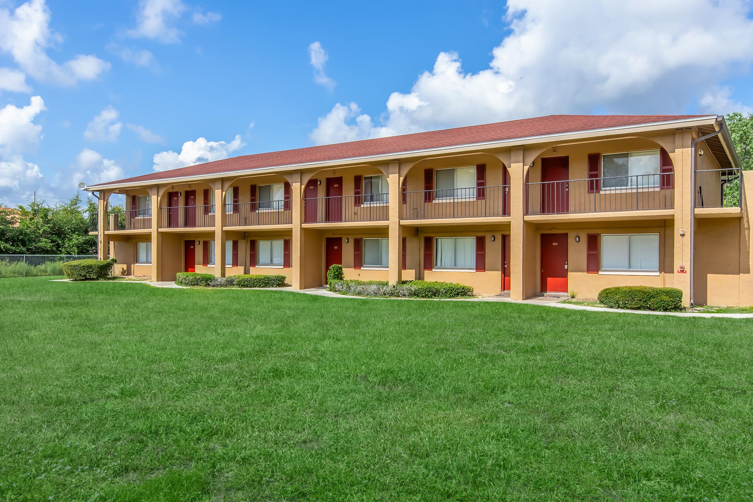 A two-story apartment building with a beige exterior and red doors, set against a blue sky with fluffy white clouds. The lawn is green and well-maintained, with small shrubs lining the building's front. The building features balconies and a simple architectural style.