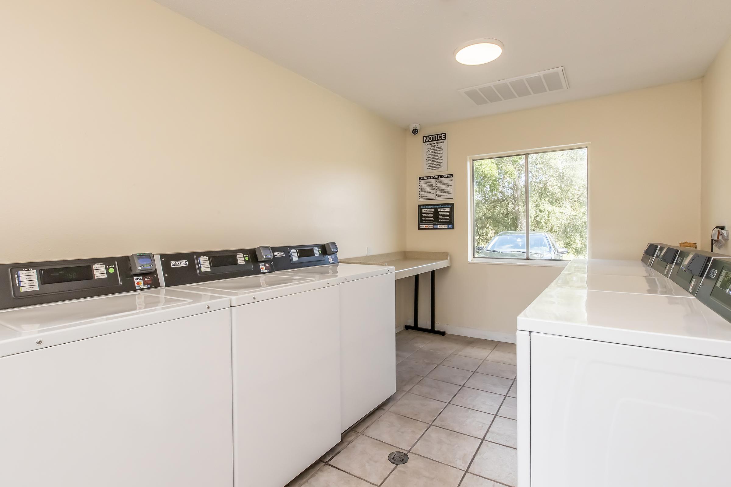 A clean, well-lit laundry room featuring several white washing machines and dryers lined up against the wall. There is a table for folding clothes and a window with natural light coming in. A poster with laundry instructions is displayed on the wall. The floor is tiled.
