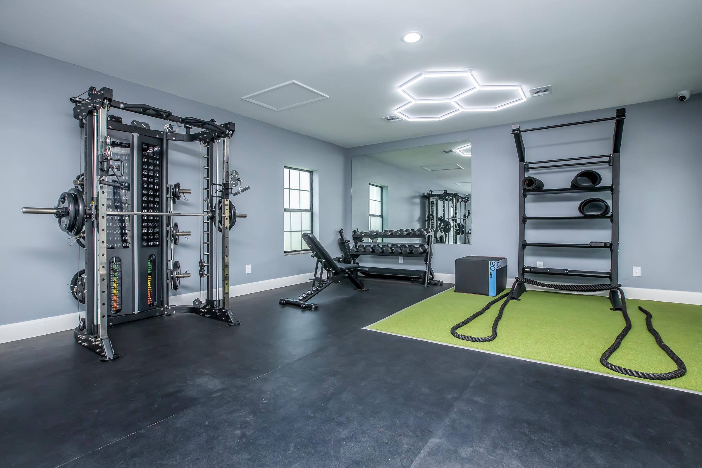 A modern home gym featuring a multi-station weight machine, adjustable workout bench, a large mirror, and storage for free weights. The floor is rubber with a green turf area, and there are battle ropes neatly coiled. Bright lighting and a minimalist design create an inviting workout space.
