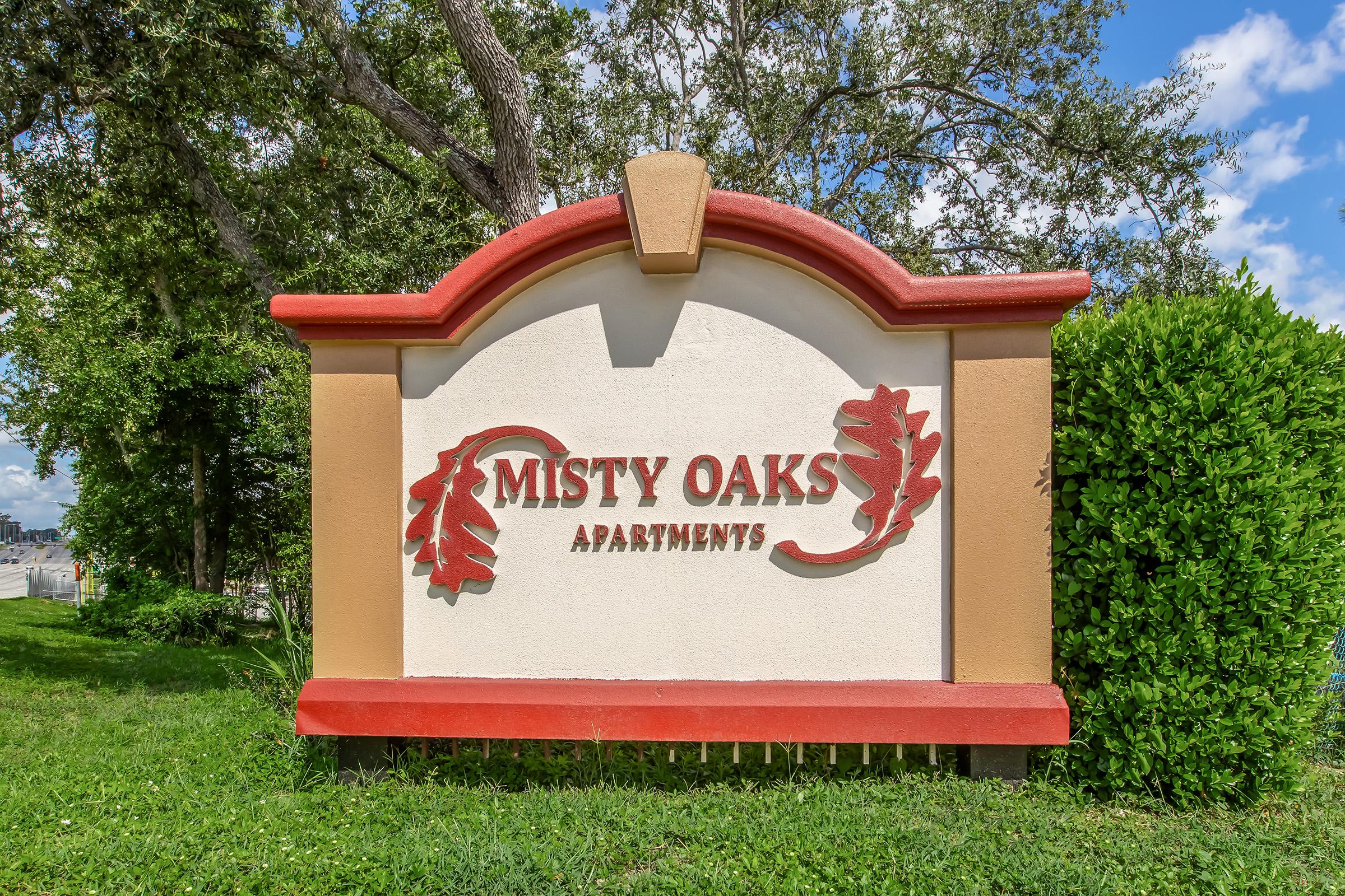 Sign for Misty Oaks Apartments featuring decorative leaf motifs, set against a backdrop of green foliage and blue sky. The sign is prominently displayed with bold, red lettering on a lighter beige background.