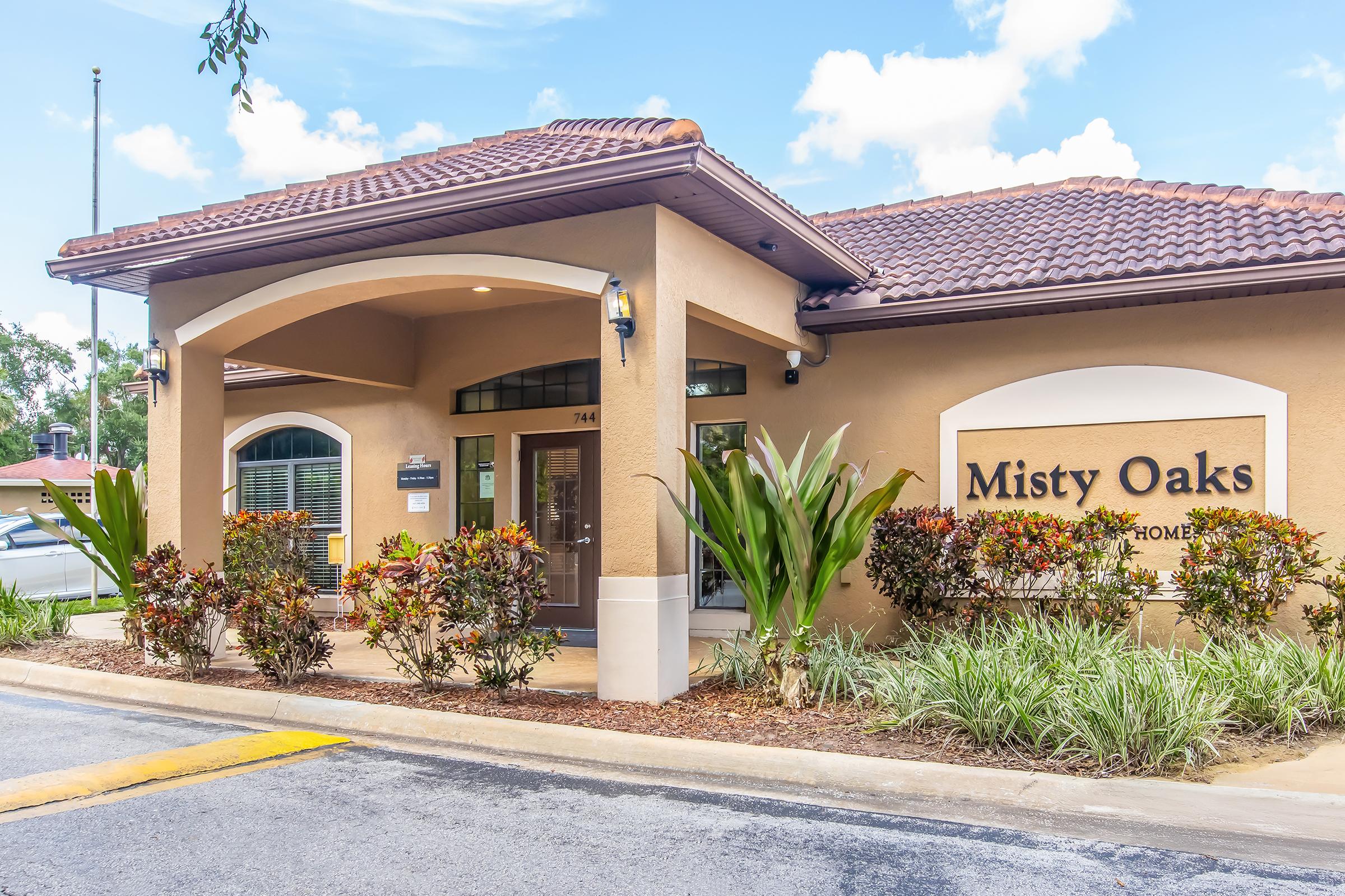 Exterior view of the Misty Oaks residential community entrance, featuring a welcoming facade with a brown roof, landscaped shrubs, and decorative plants. A sign with the name "Misty Oaks" is prominently displayed near the entrance, which has large windows and a glazed door. The setting includes a clear blue sky with a few clouds.