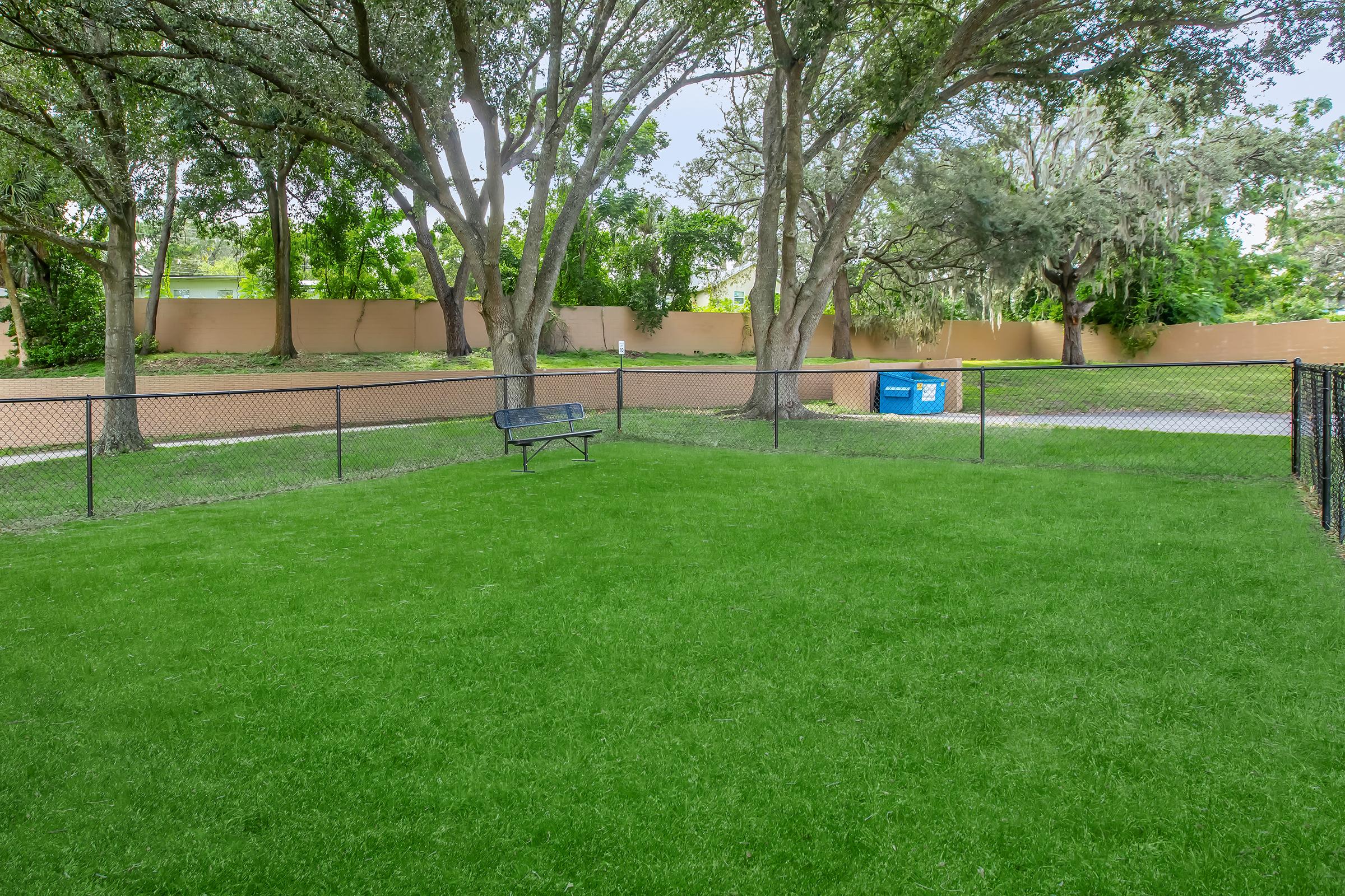 A green grassy area enclosed by a black fence, featuring a single bench on the lawn. In the background are two large trees and a dumpster near a wall. The area appears well-maintained and is likely part of a park or recreational space.