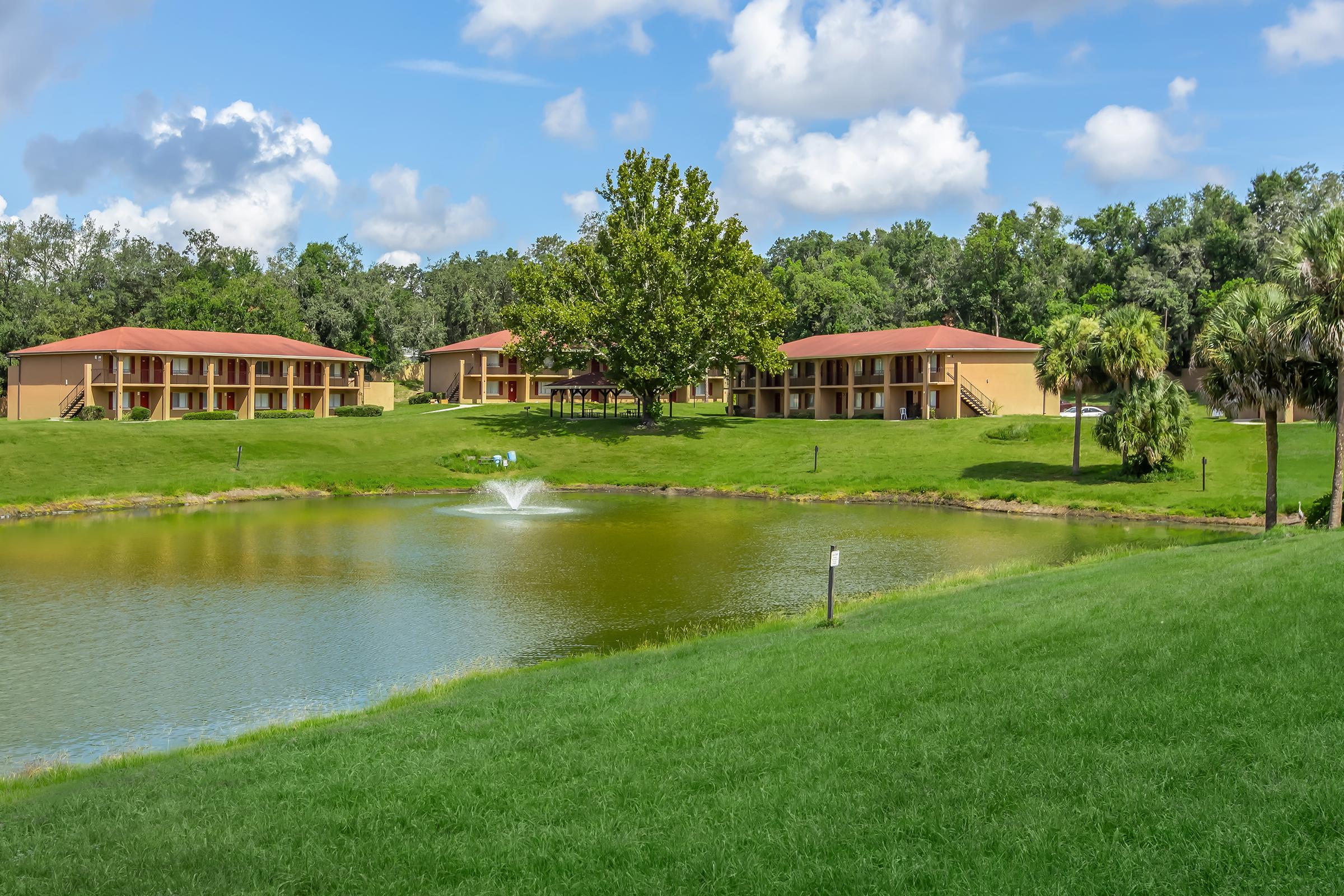 A scenic view of several low-rise buildings beside a calm pond, featuring a fountain in the water. Lush green grass and trees surround the area, under a partly cloudy blue sky. The setting appears tranquil and inviting, ideal for a relaxed outdoor atmosphere.
