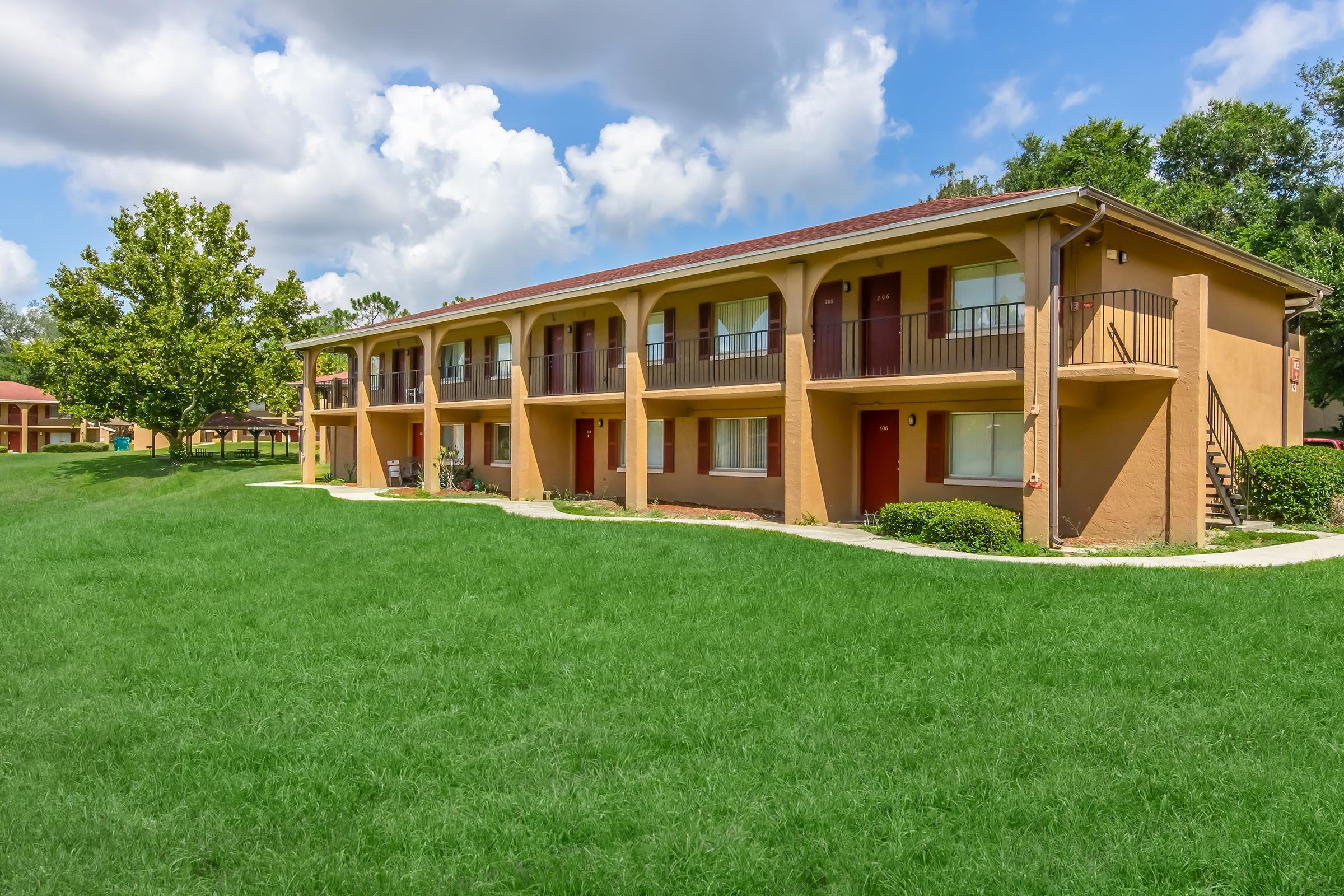 A two-story building with a brown exterior, featuring multiple balconies and doors. The structure is surrounded by lush green grass and trees, under a partly cloudy sky. There are sidewalks leading to the entrances, suggesting a well-maintained outdoor space.