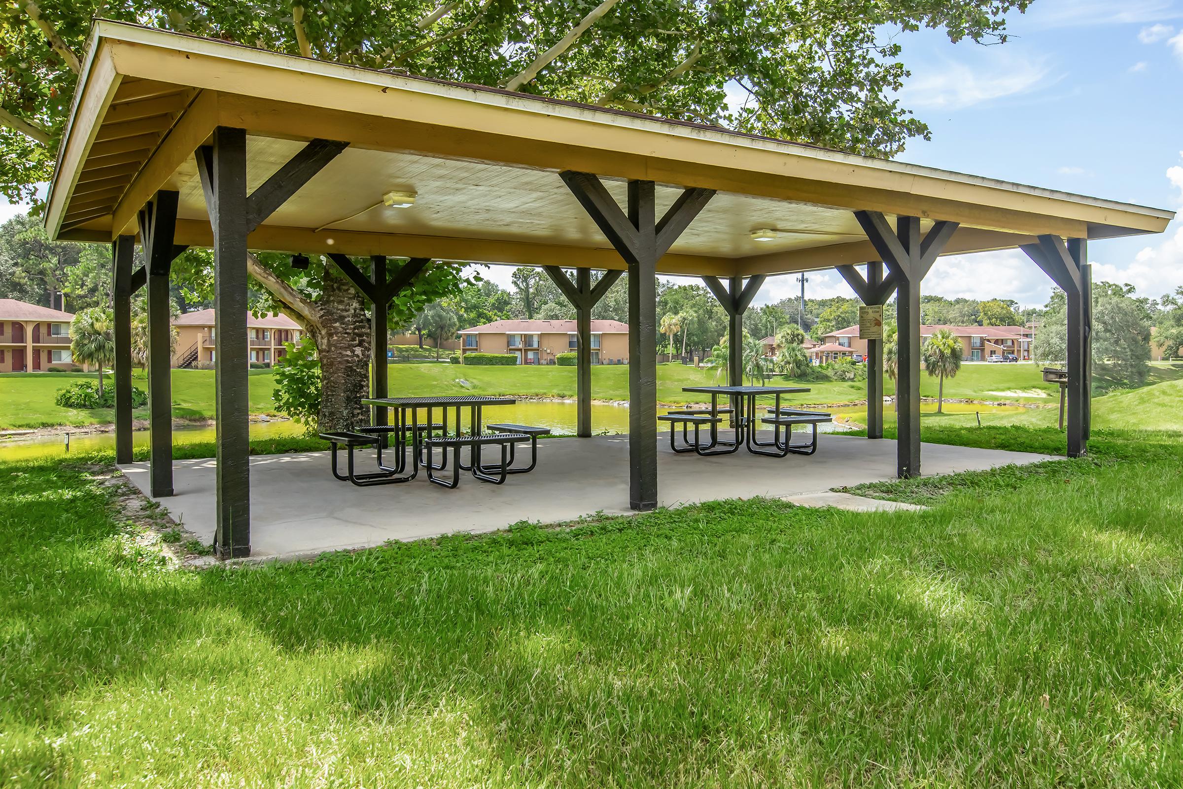 A shaded picnic area with a wooden pavilion supported by black posts, featuring several picnic tables. The setting includes green grass and a tranquil pond in the background, surrounded by residential buildings and trees under a clear blue sky.