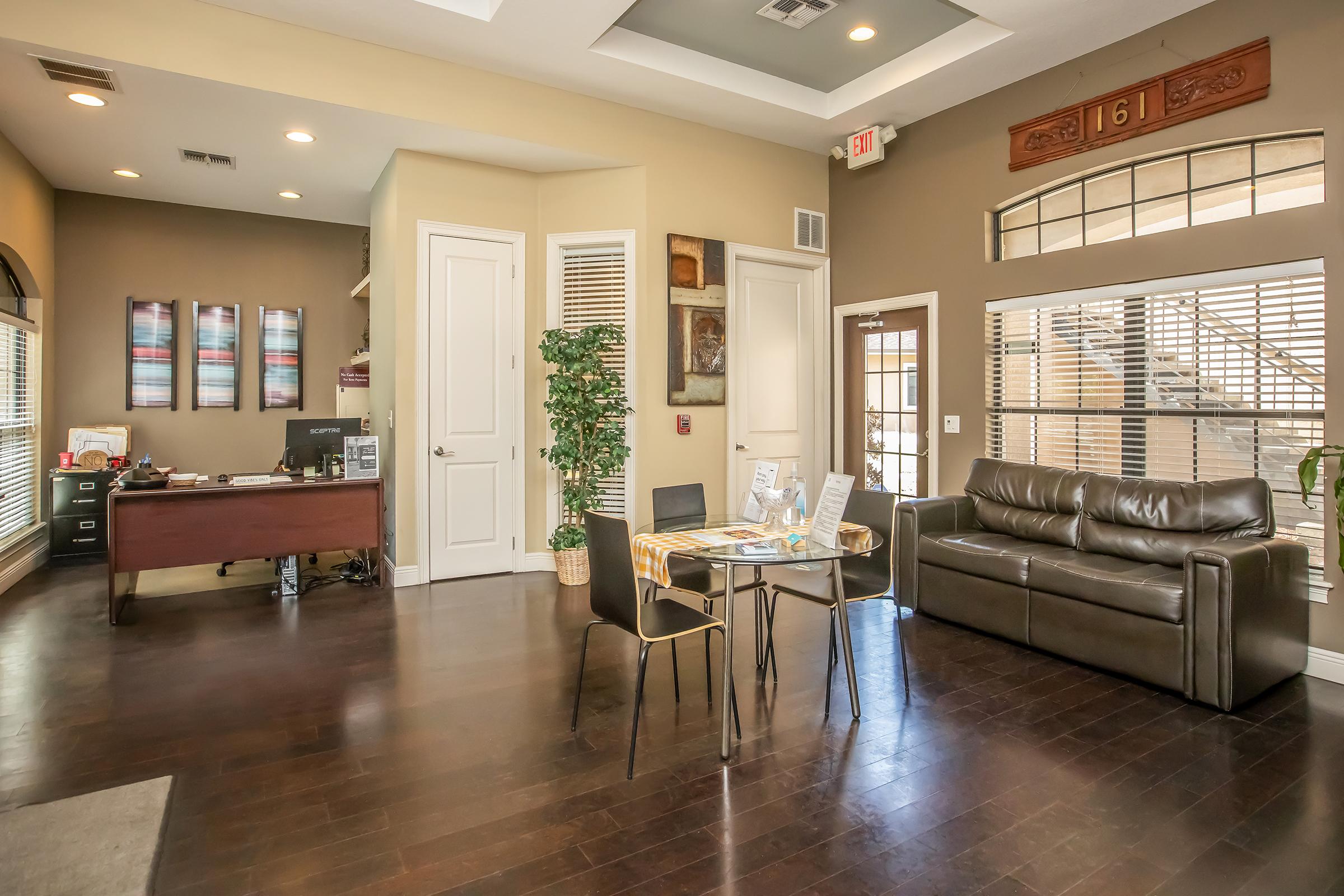 An inviting office reception area featuring a desk with a computer, a cozy brown leather sofa, and a small dining table with chairs. The walls are painted in soft tones, adorned with artwork, while natural light comes through large windows. A potted plant adds a touch of greenery.