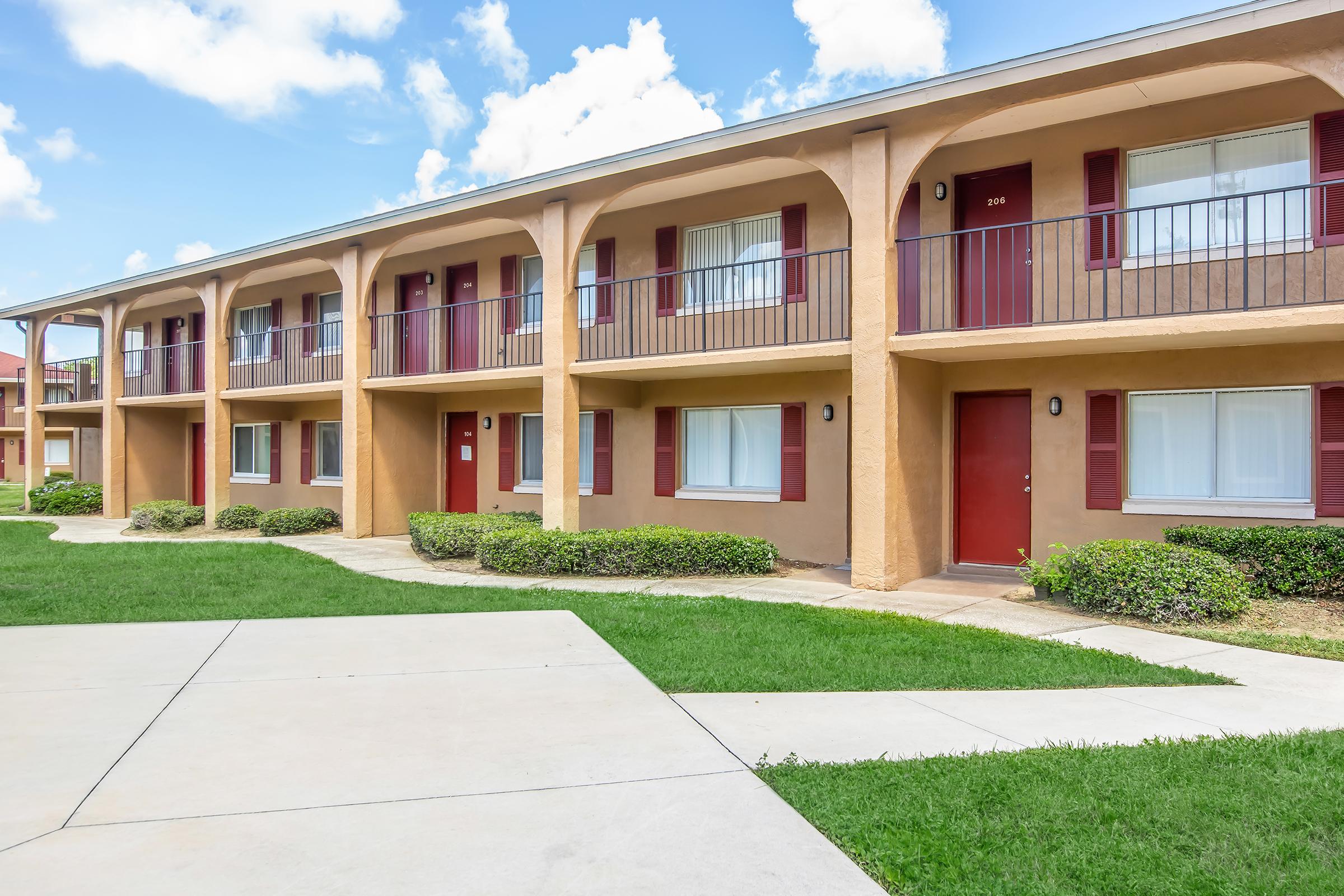 A two-story apartment building with a sandy beige exterior and red shutters. The building features several balconies with railings. A well-maintained grassy area surrounds the building, and a concrete pathway leads to the entrance doors, which are visibly numbered. The sky is bright with scattered clouds.