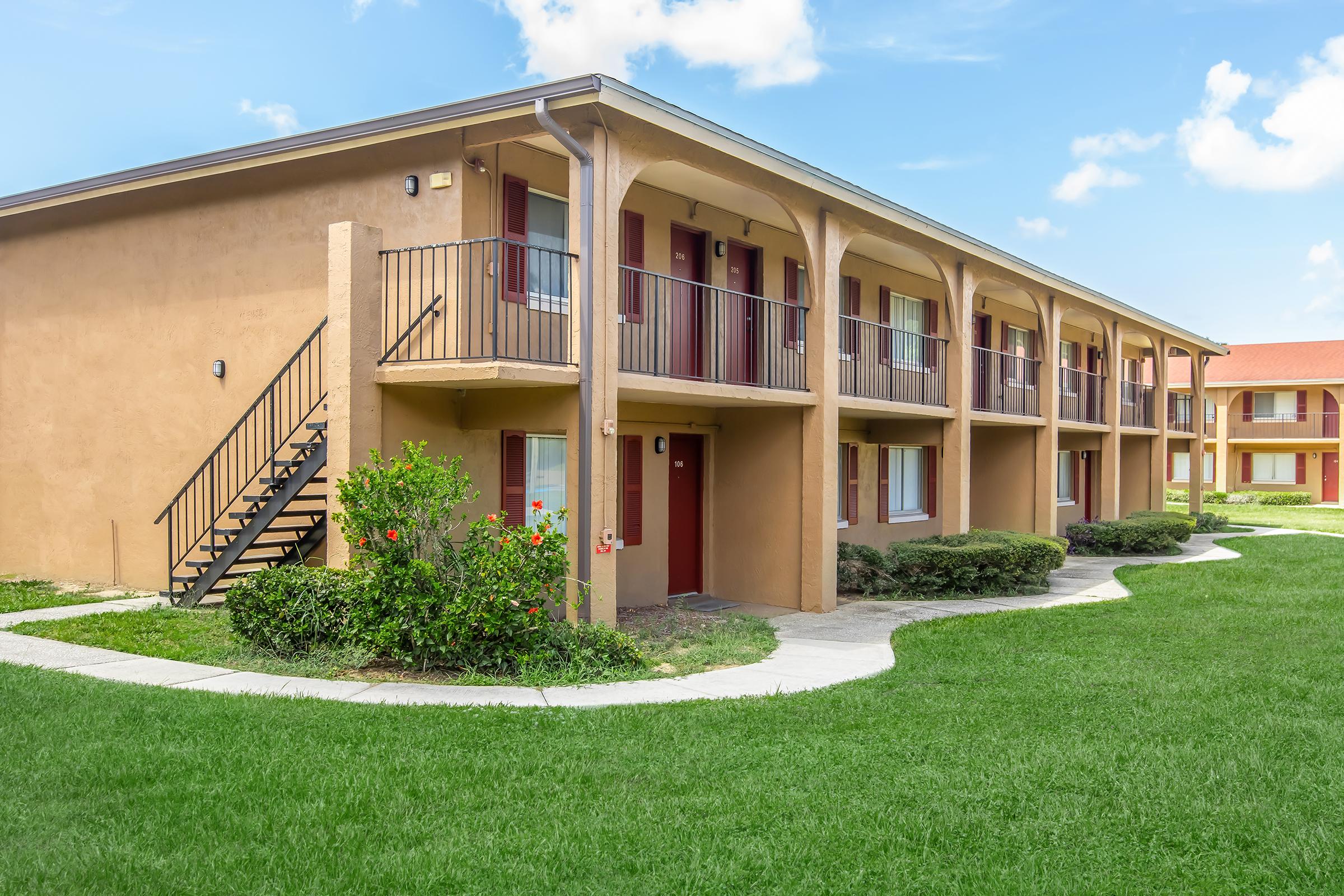 Two-story building featuring several balcony-style rooms with red doors, surrounded by well-maintained green lawns and shrubbery. A walkway leads to the entrance, and a staircase provides access to the upper floor. The sky is clear with a few clouds visible.