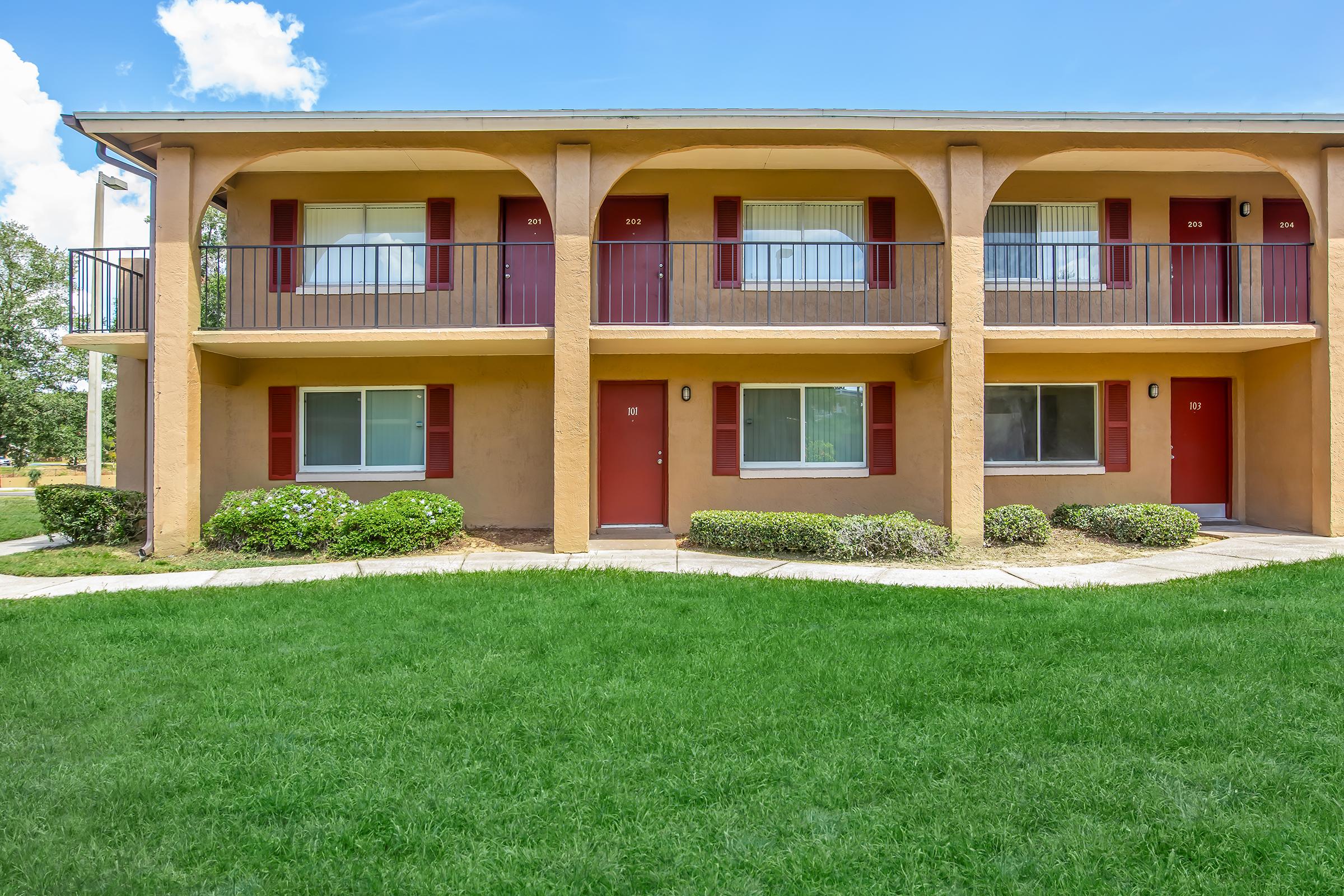 Two-story apartment building with an orange exterior and multiple entrances. Each unit has red doors and white shutters. The building is surrounded by a well-maintained green lawn and shrubs, with a clear blue sky above. The architectural style features arched balconies.