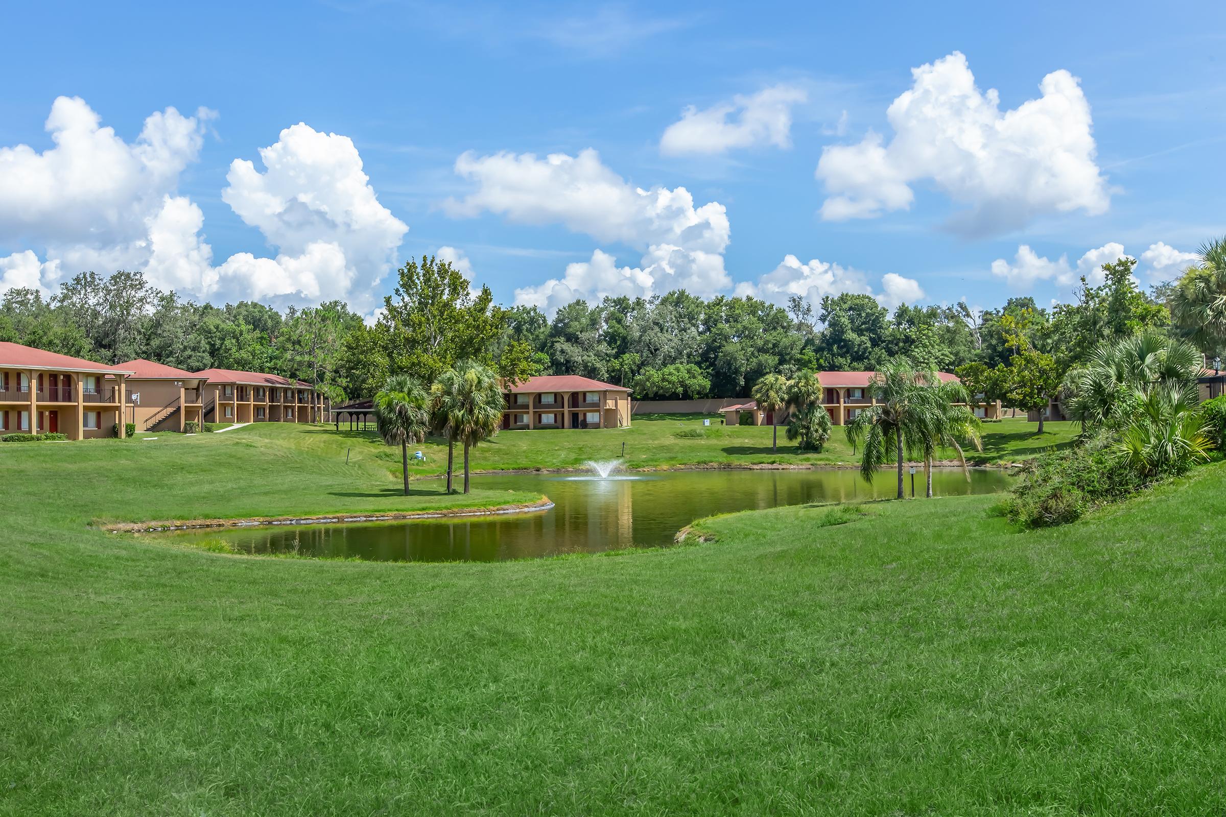 A scenic view of a landscaped area featuring a calm pond with a small fountain, surrounded by lush green grass and palm trees. In the background, there are several residential buildings with red roofs, set against a backdrop of blue skies and fluffy white clouds.