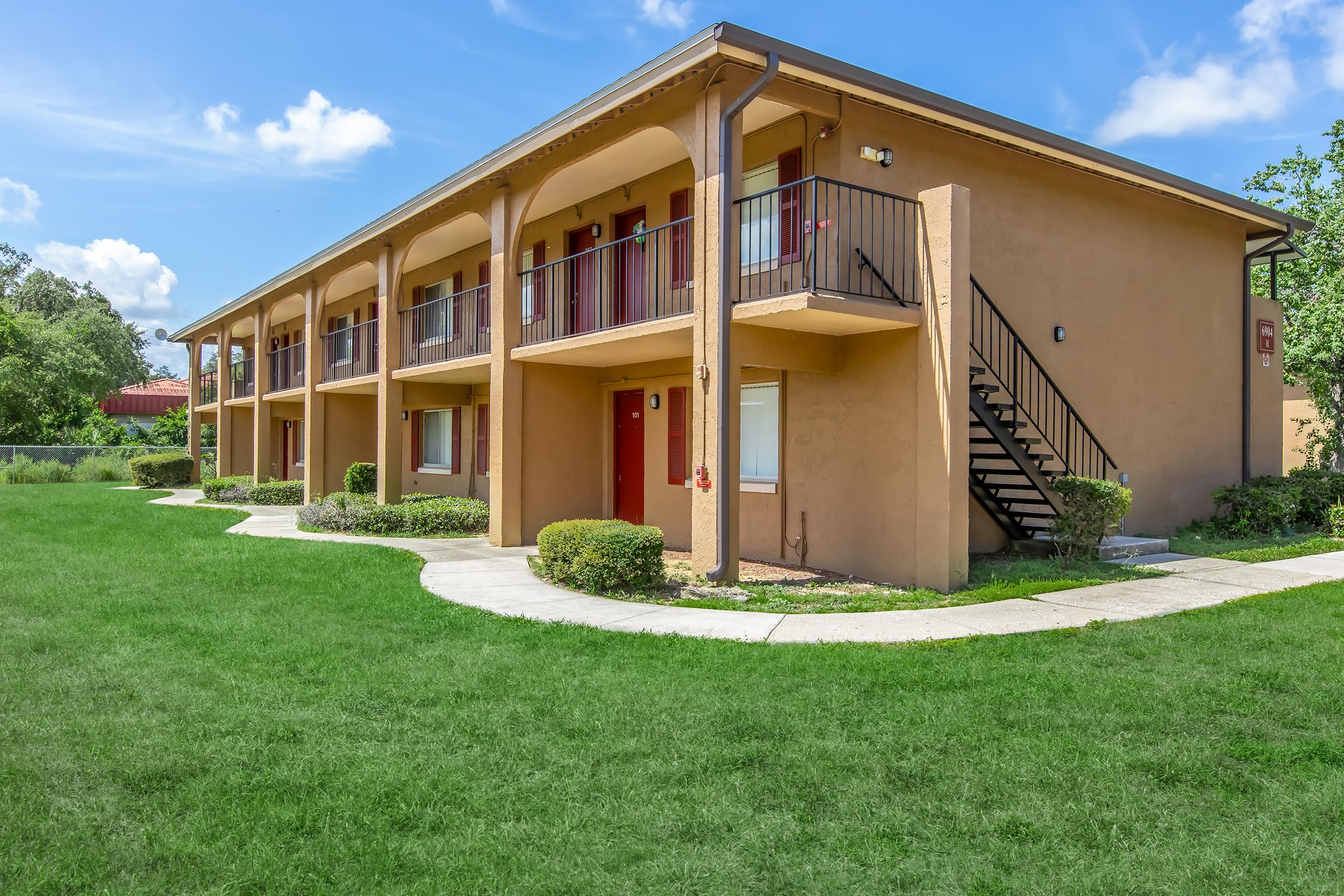 A two-story building featuring several balconies and red doors, surrounded by a well-maintained lawn and shrubs. The exterior is painted in a warm beige tone, with stairs leading to the upper level. Clear blue skies and scattered clouds are visible in the background.