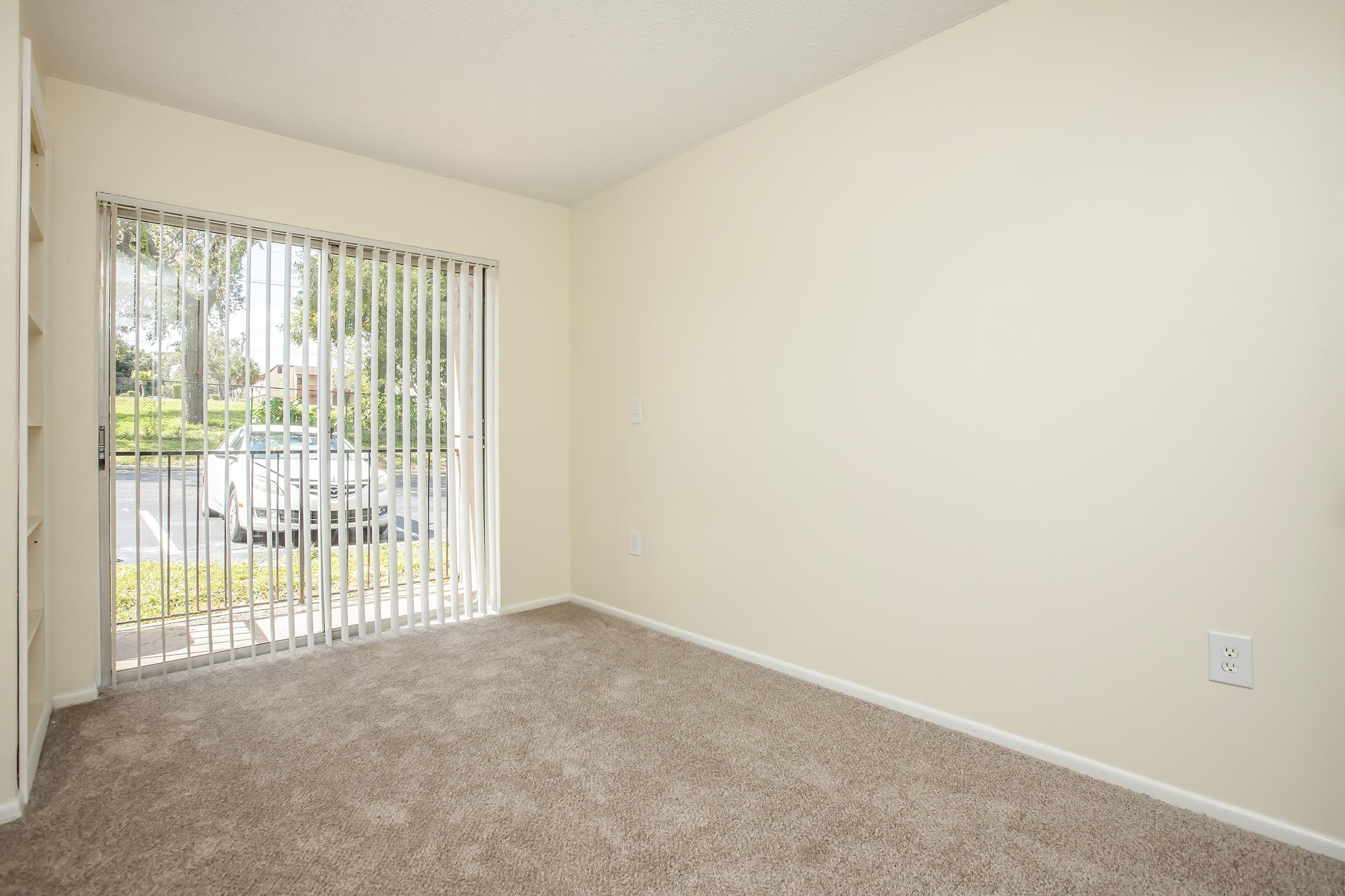 A vacant room with beige walls and light brown carpet. The room features one large window with vertical blinds, partially open, showing a view of greenery and parked cars outside. There are shelves built into one wall, creating a simple, uncluttered space.