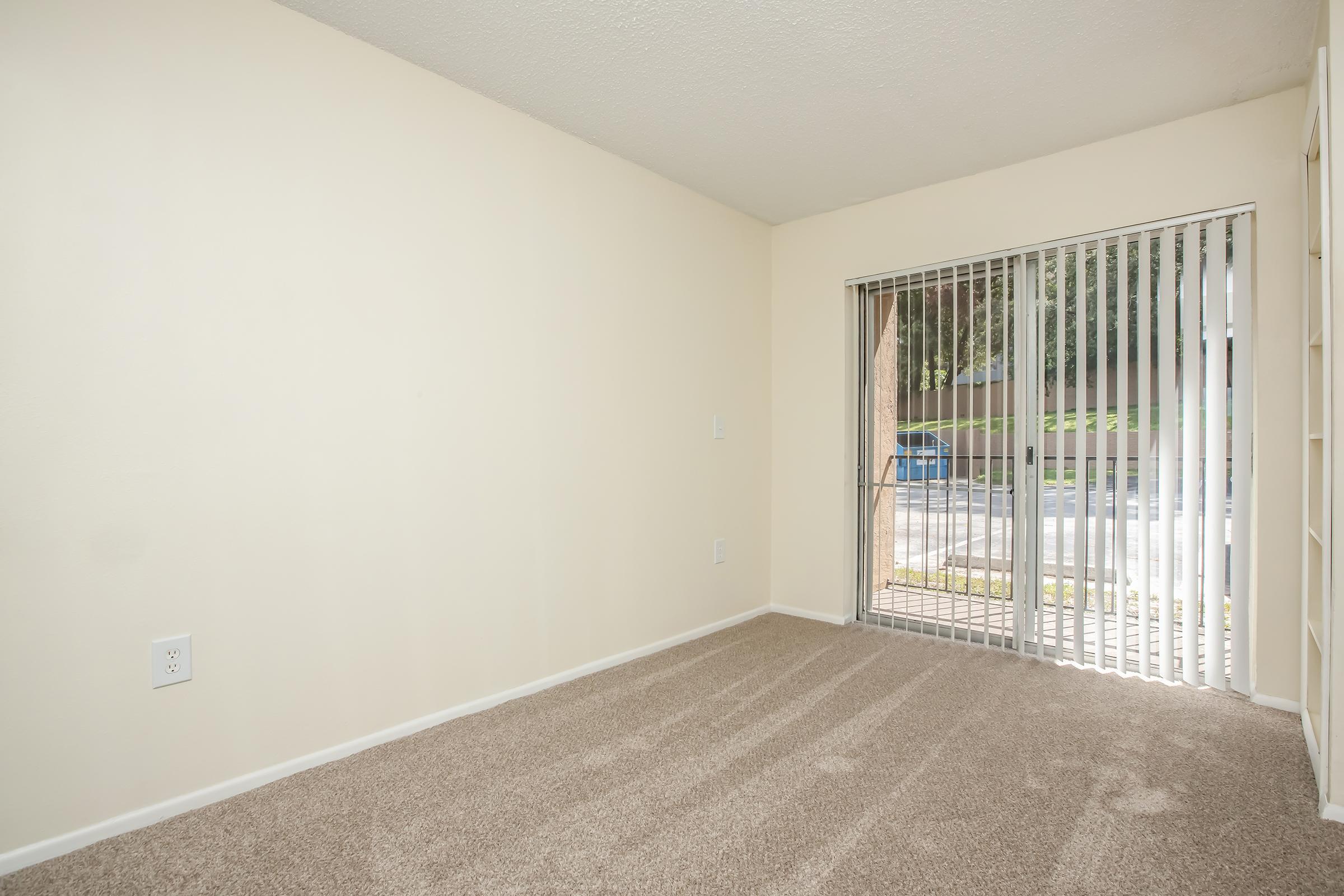 Empty room with beige walls and carpeted flooring. A large sliding glass door with vertical blinds lets in natural light, revealing a view of outdoor greenery. The space is uncluttered, with no furniture or decorations present, creating a versatile area ready for personalization.