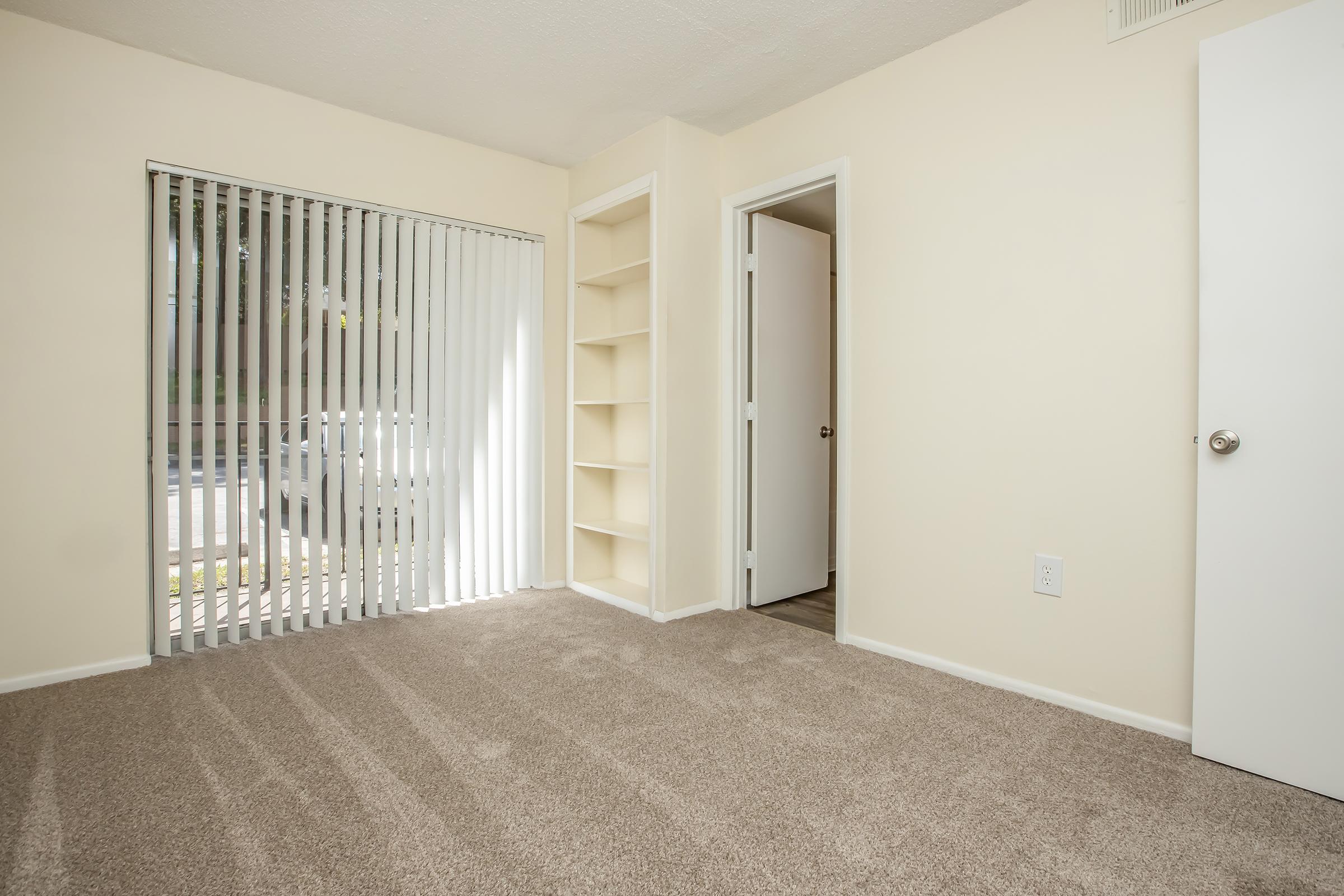 An empty room featuring light beige walls and carpeted flooring. There are vertical blinds covering a large window, allowing natural light to enter. To the right, there is an open door leading to a hallway, and a built-in shelf unit against the left wall.