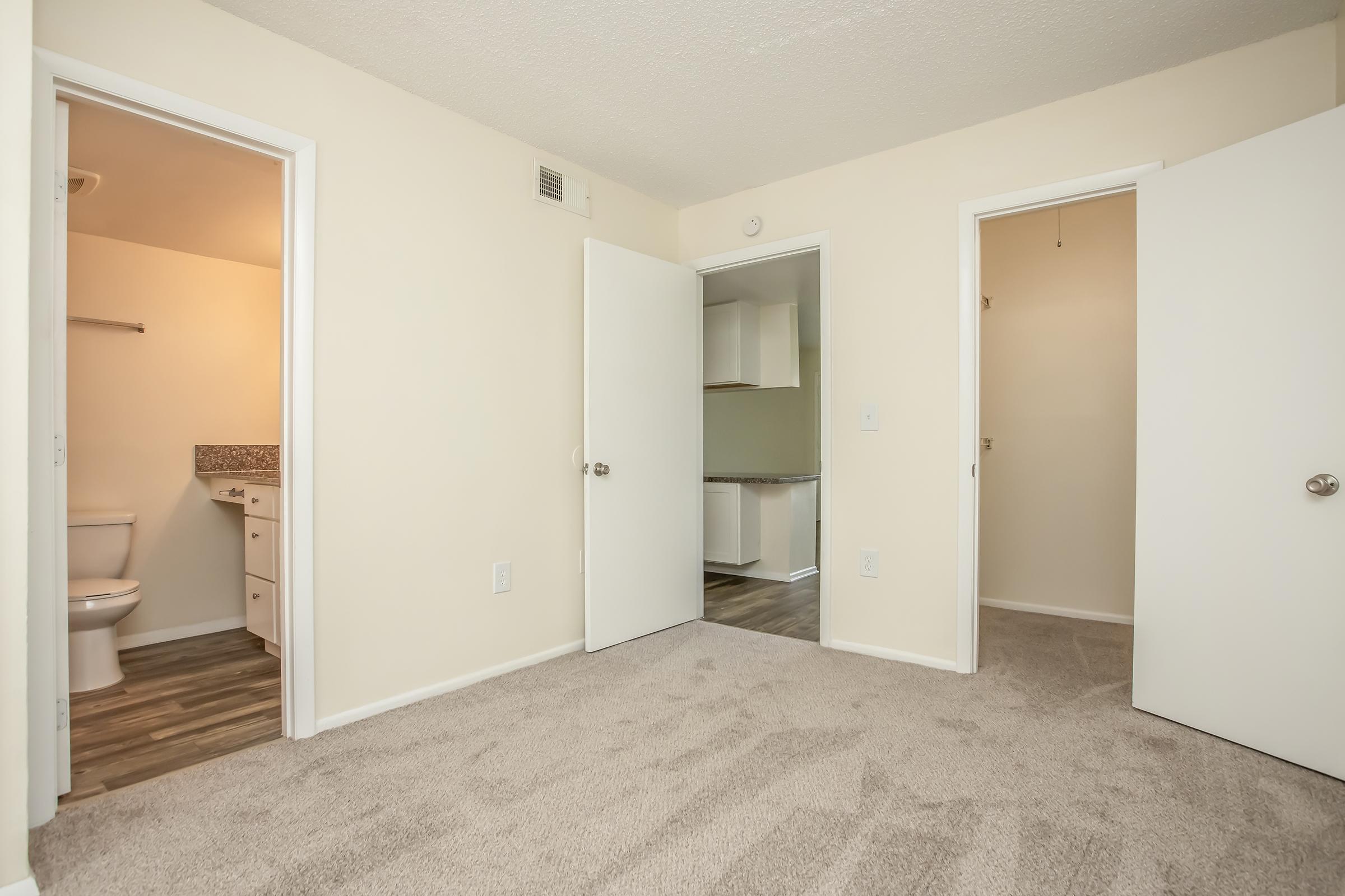 Interior view of a light-colored room with beige carpet, featuring two open doors; one leads to a bathroom with a toilet and sink, and the other opens to a kitchen area. The walls are painted in neutral tones, creating a spacious and inviting atmosphere.