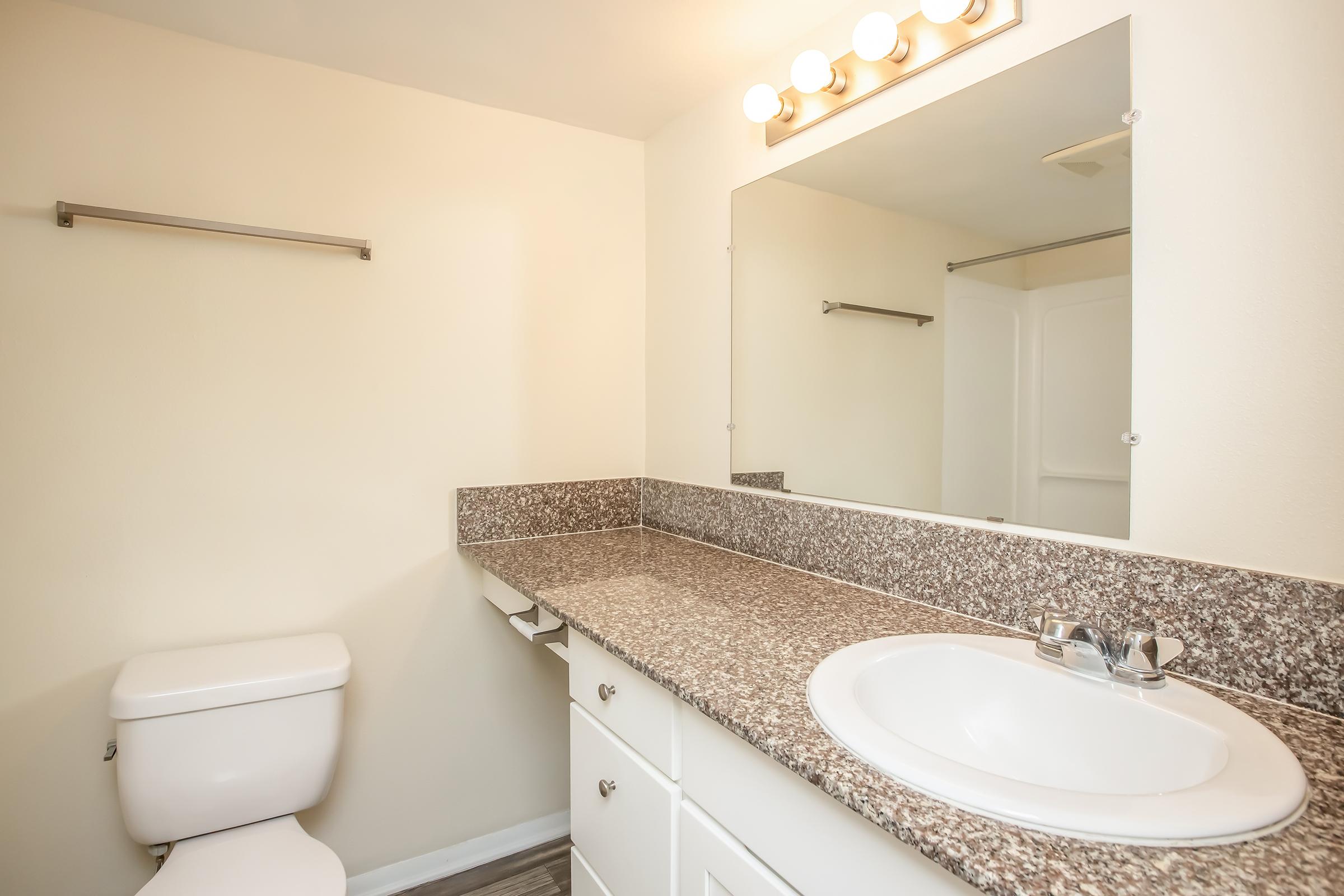 A clean and modern bathroom featuring a white toilet, a countertop with a sink, and a large mirror above the sink. The wall is painted a light color, and there's a towel bar next to the mirror. The flooring appears to be gray.
