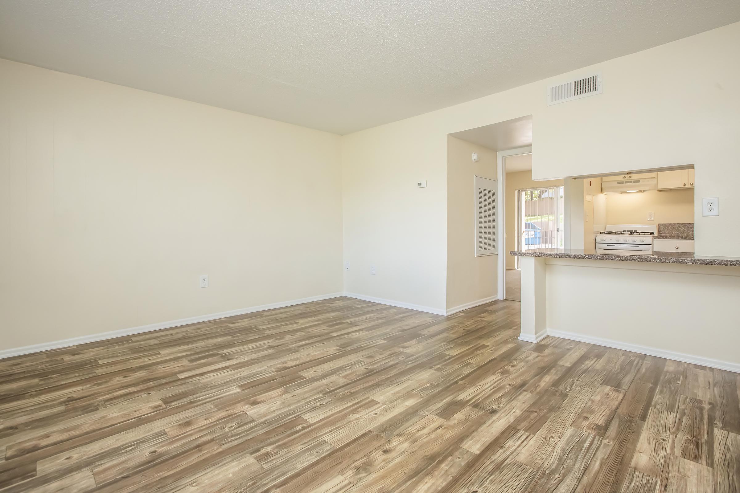 A spacious, empty living room with light-colored walls and laminate flooring. A doorway on the right leads to a kitchen area with white appliances visible through a cutout window. Natural light enters from a nearby window, creating a bright and inviting atmosphere.