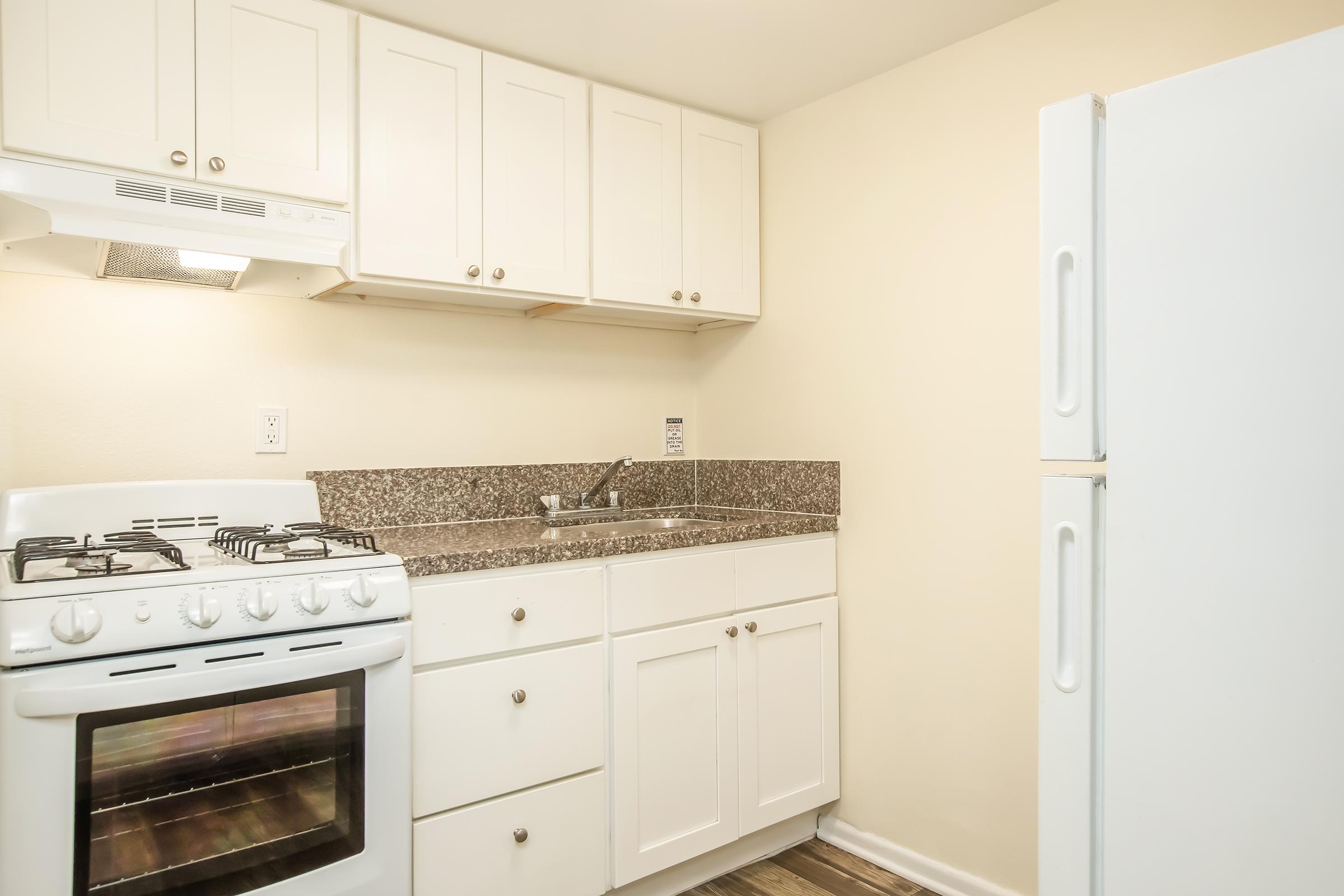 A small, modern kitchen with white cabinetry, a granite countertop, a gas stove with an overhead vent, a stainless steel sink, and a white refrigerator. The walls are light-colored, and the flooring is a wood-like texture. The space appears clean and well-maintained.