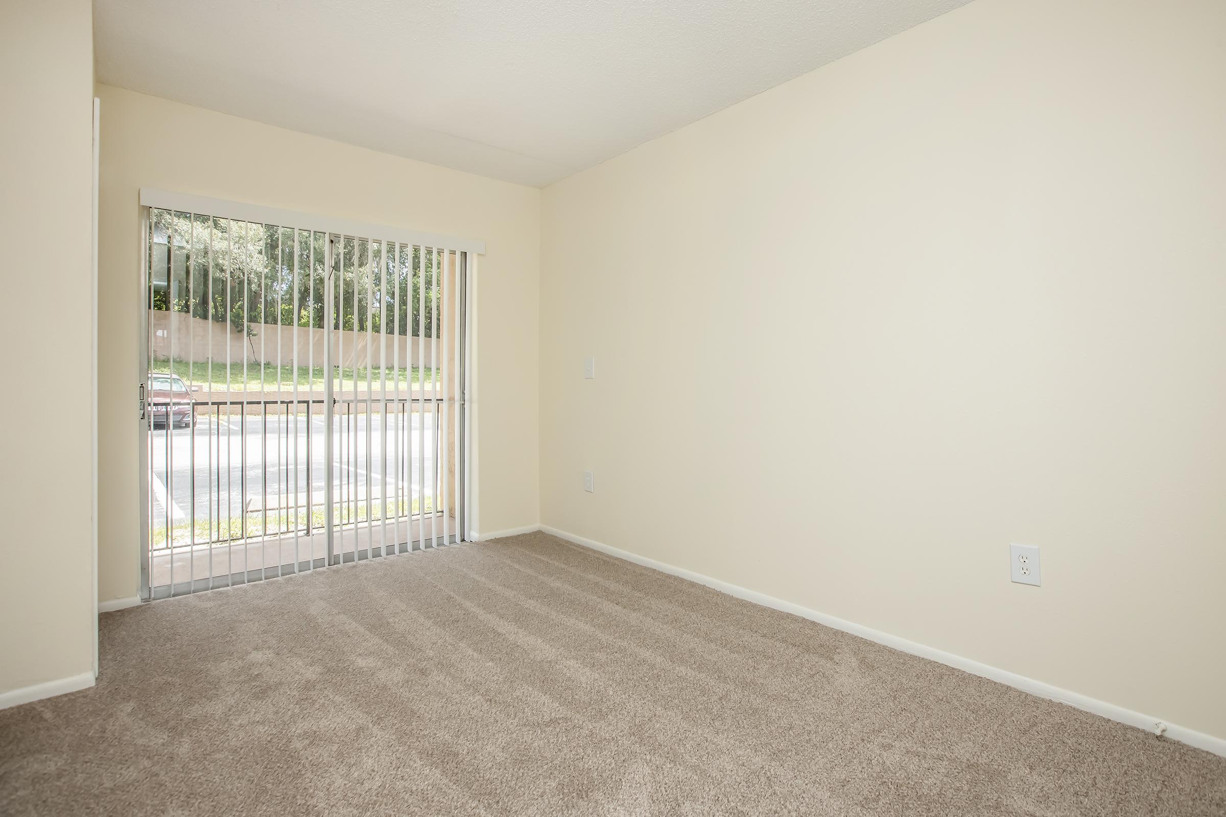 A small, empty room with beige walls and carpet. A sliding glass door with vertical blinds leads to an outdoor area. Natural light filters in from the outside, illuminating the space. The room has no furniture, creating a clean and open feel.