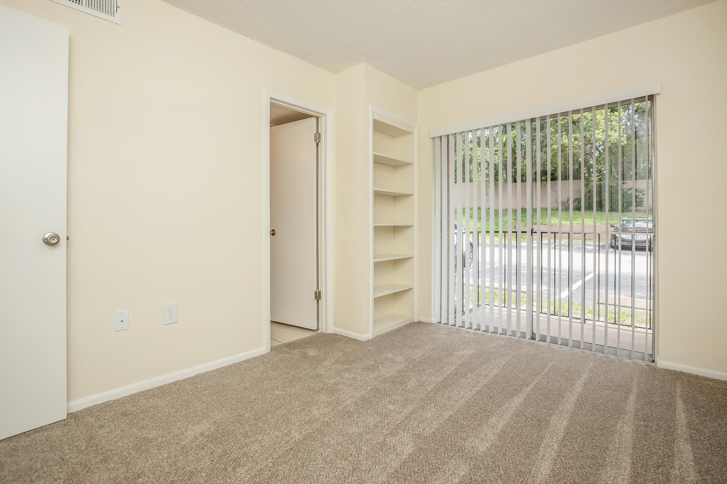 A carpeted room with beige walls and a large window featuring vertical blinds. There is a door on the left leading to another room, and a built-in shelf unit on the right. The window overlooks a parking area outside, with greenery visible in the background.
