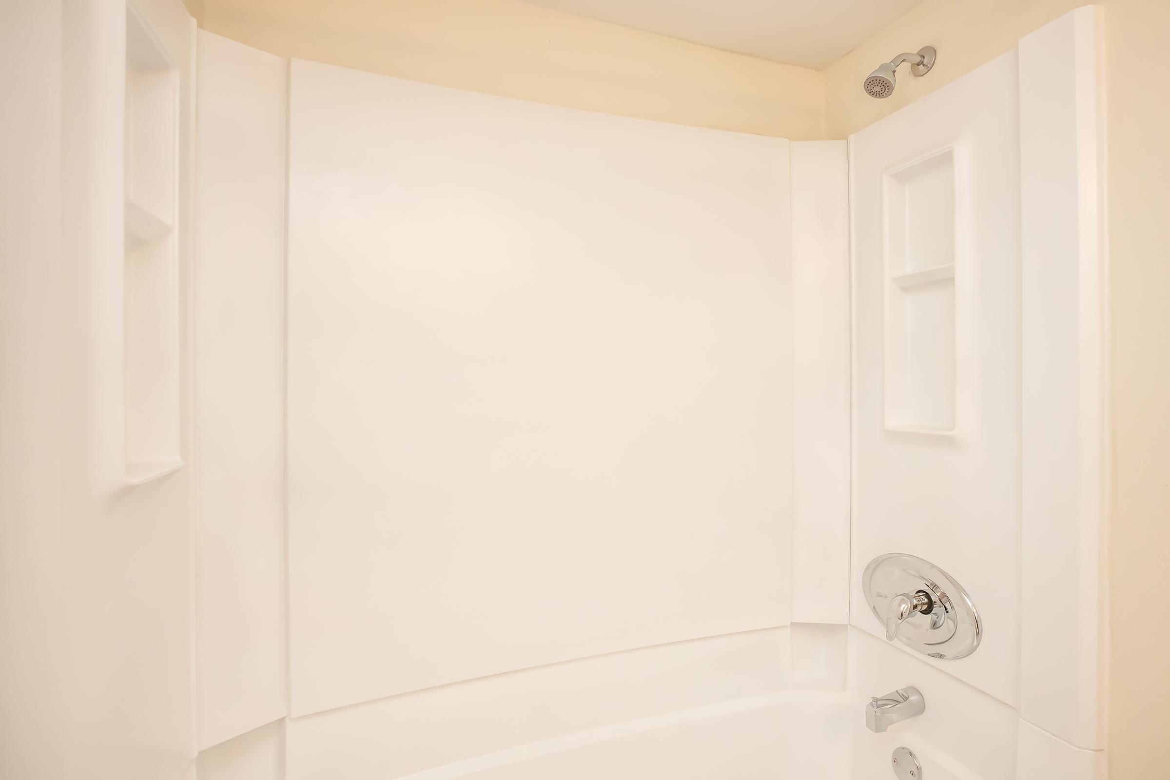 A clean, modern shower stall featuring smooth white walls, a showerhead on the right, and built-in shelves for toiletries. The overall design is minimalistic, with a curved wall providing an open feel to the space.