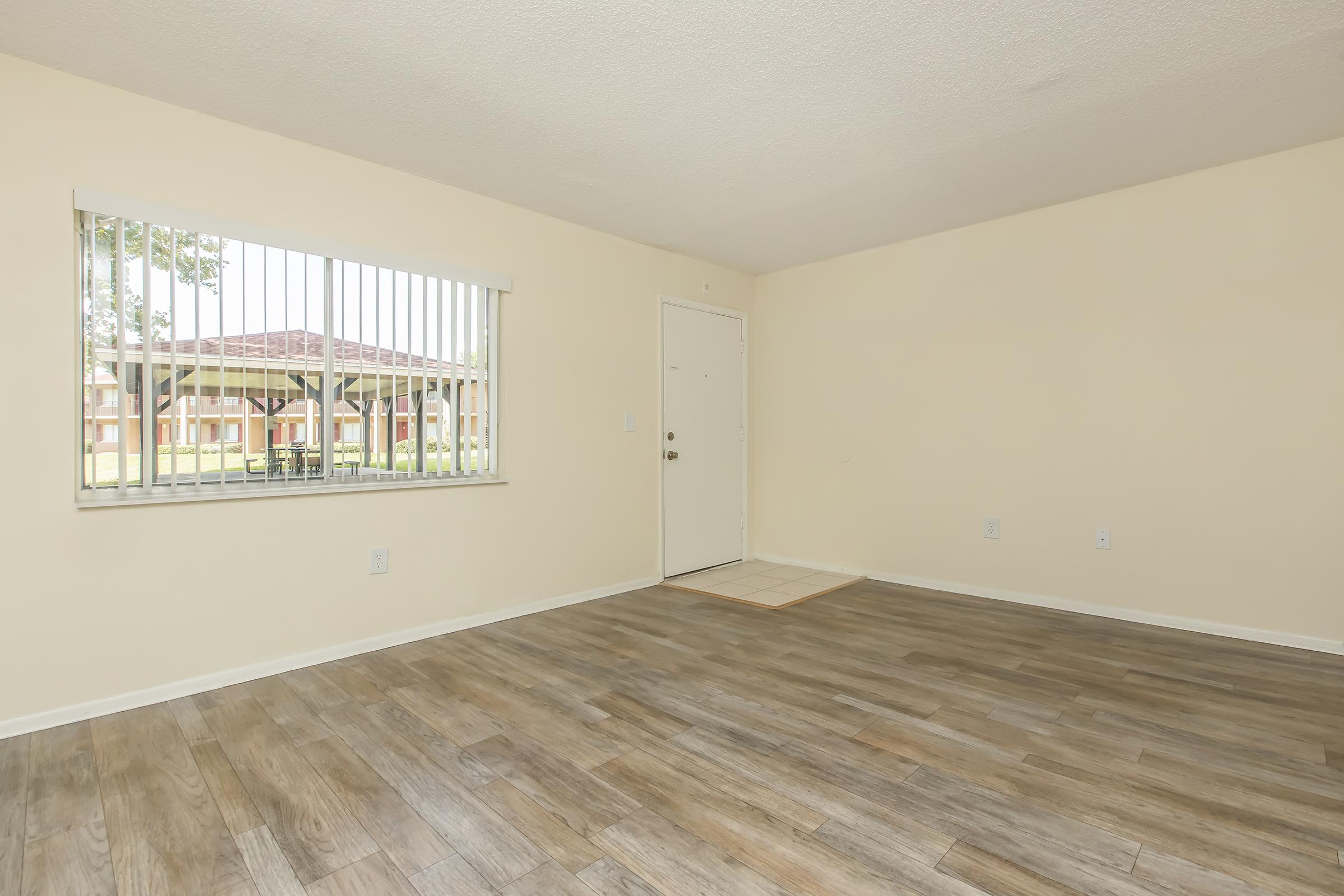 A spacious, empty room with light beige walls and wooden flooring. It features a large window with white blinds, allowing natural light to enter. The door to the outside is visible, and the overall space is clean and uncluttered, ideal for various furniture arrangements.