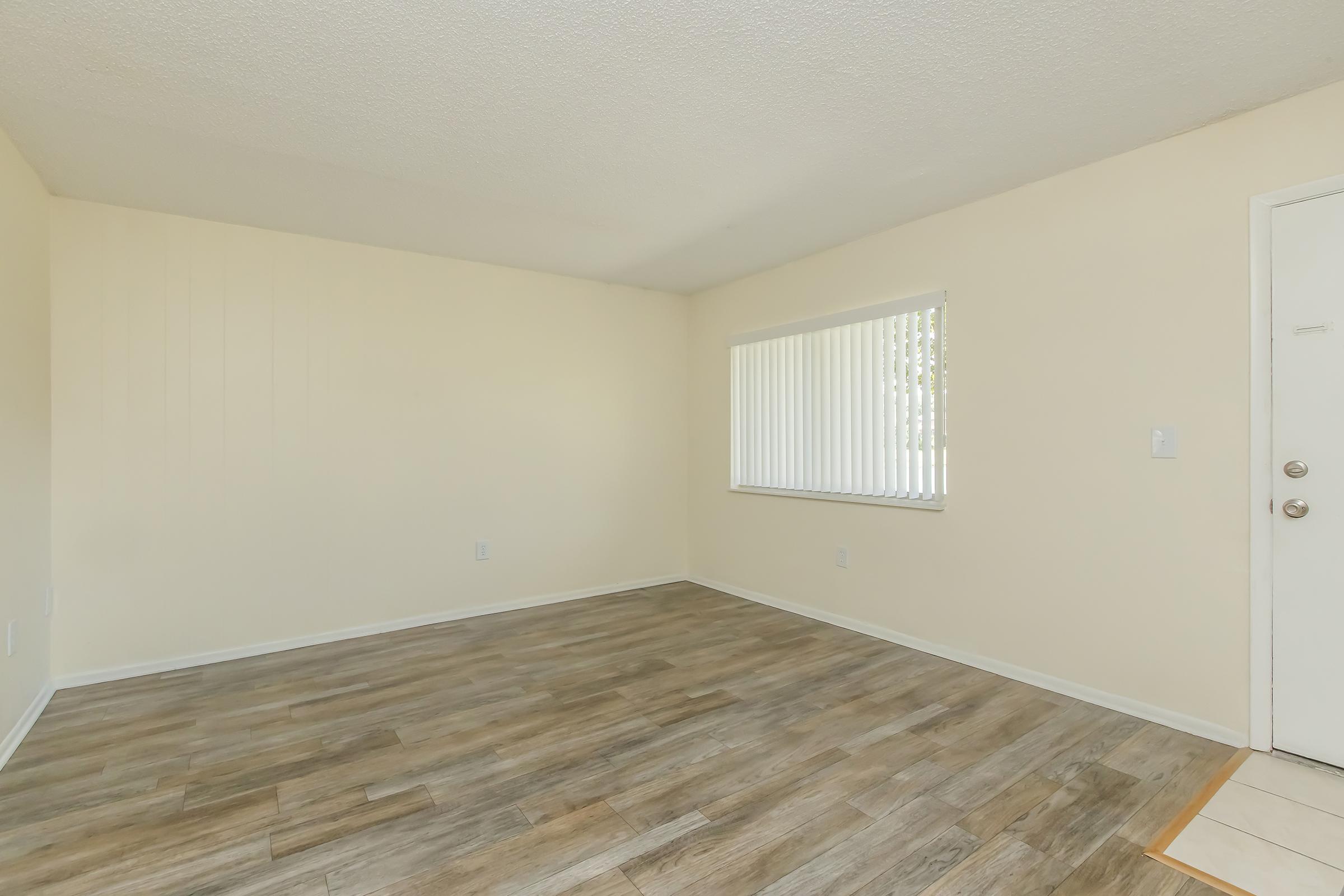 A spacious, empty living room with beige walls and wooden flooring. There is a large window with vertical blinds allowing natural light to enter, and a white door on the right side of the image. The overall ambiance is bright and inviting, ideal for personalizing the space.