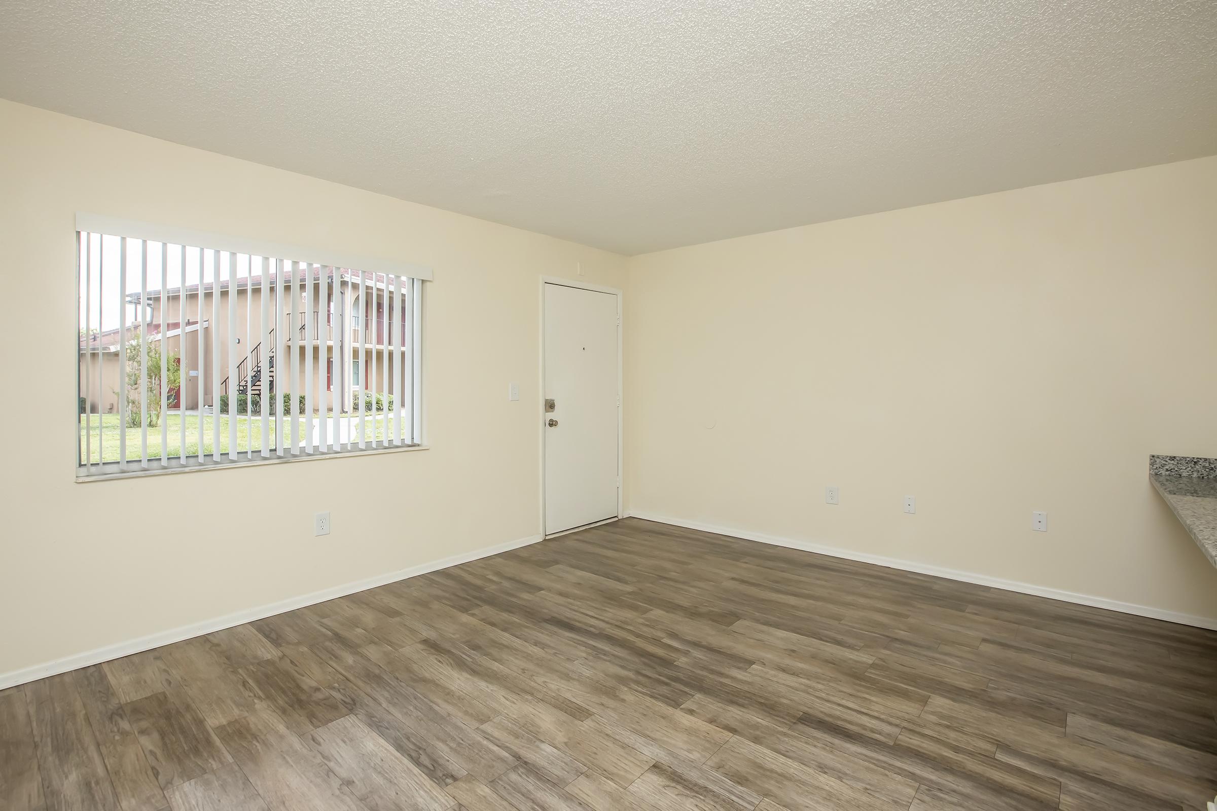 Empty room with light-colored walls and a wooden floor. A window with white blinds allows natural light in, showing greenery outside. The entrance door is visible, and there's a corner with a countertop, suggesting a kitchen area. The space appears clean and ready for furnishings.