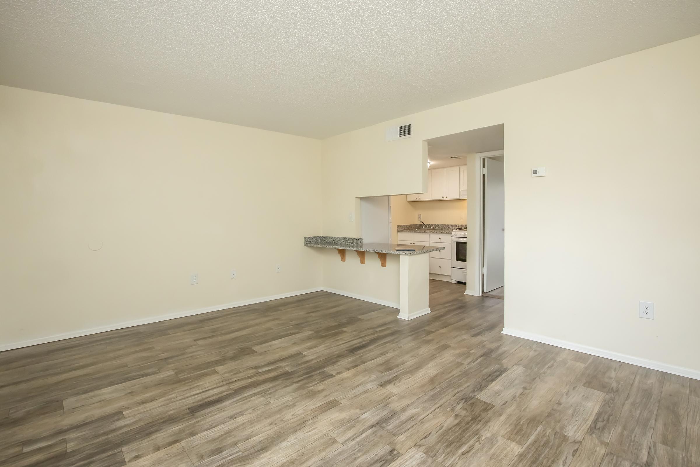 A spacious, unfurnished living area with light beige walls and laminate flooring. A small breakfast bar with a granite countertop separates the living space from a kitchen area visible in the background, featuring white cabinetry and appliances. Natural light fills the room, creating a bright atmosphere.