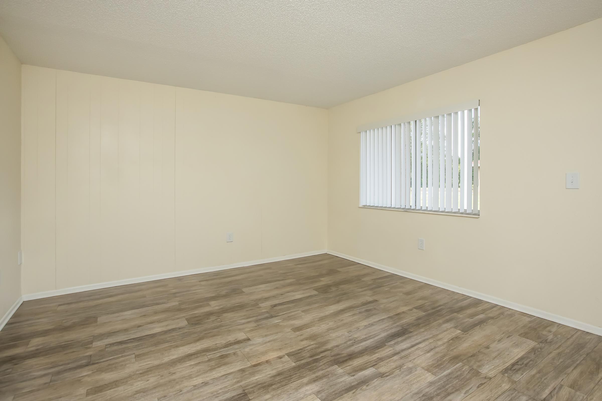 An empty room with light beige walls and a wooden floor. A set of vertical blinds covers a window that allows natural light to enter. The space is unfurnished, emphasizing its size and simplicity.